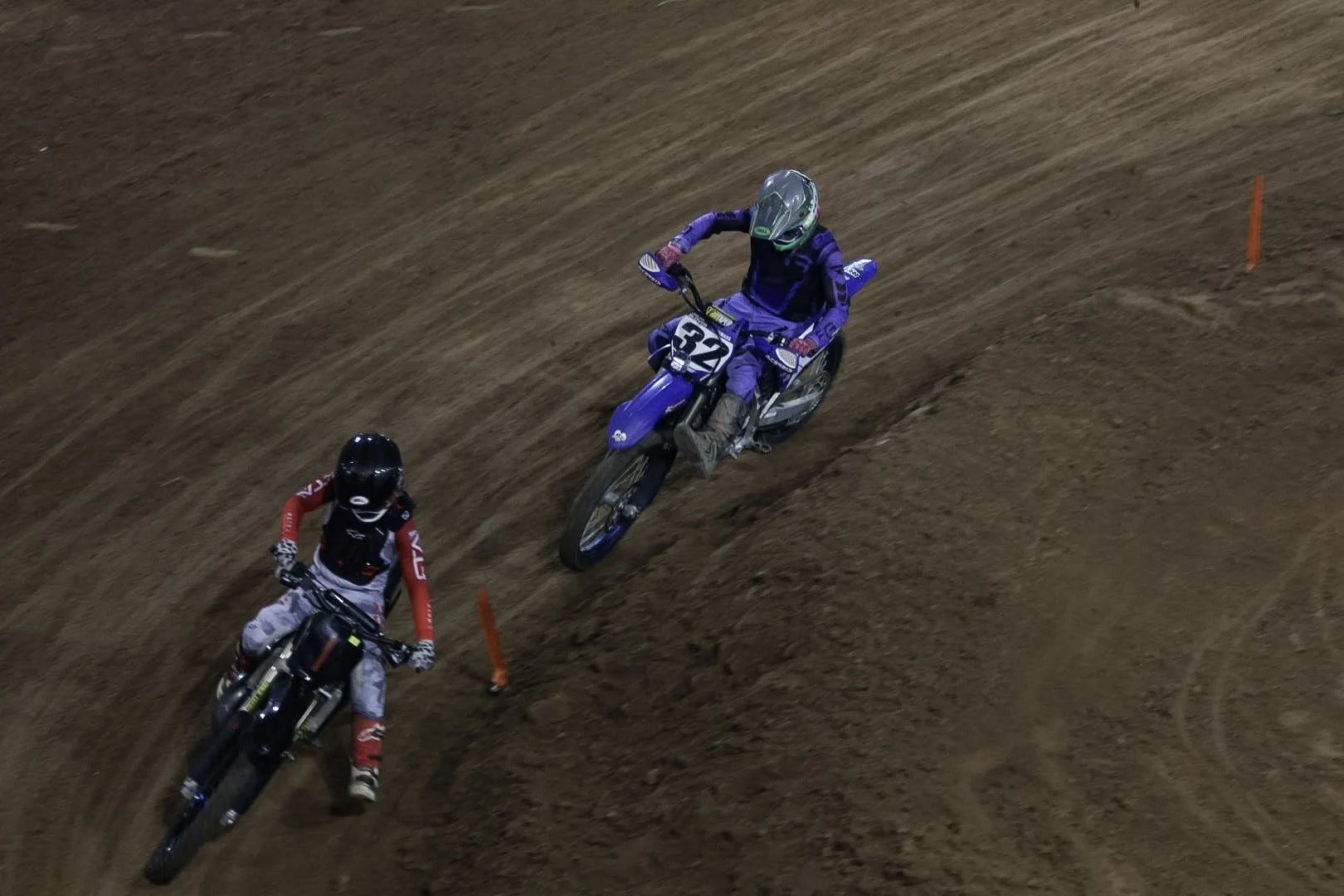 Two motocross riders racing on a dirt track, one in purple gear and the other in red and black gear.