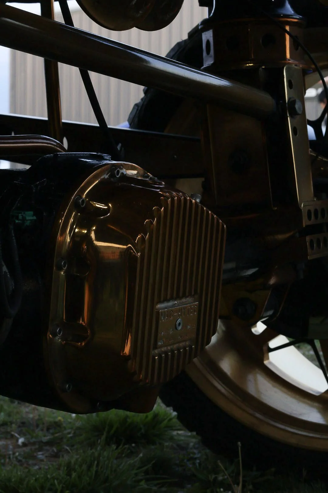 Close-up of a vintage car engine with a shiny, gold-colored component and a black frame, set outdoors with grass visible at the bottom.