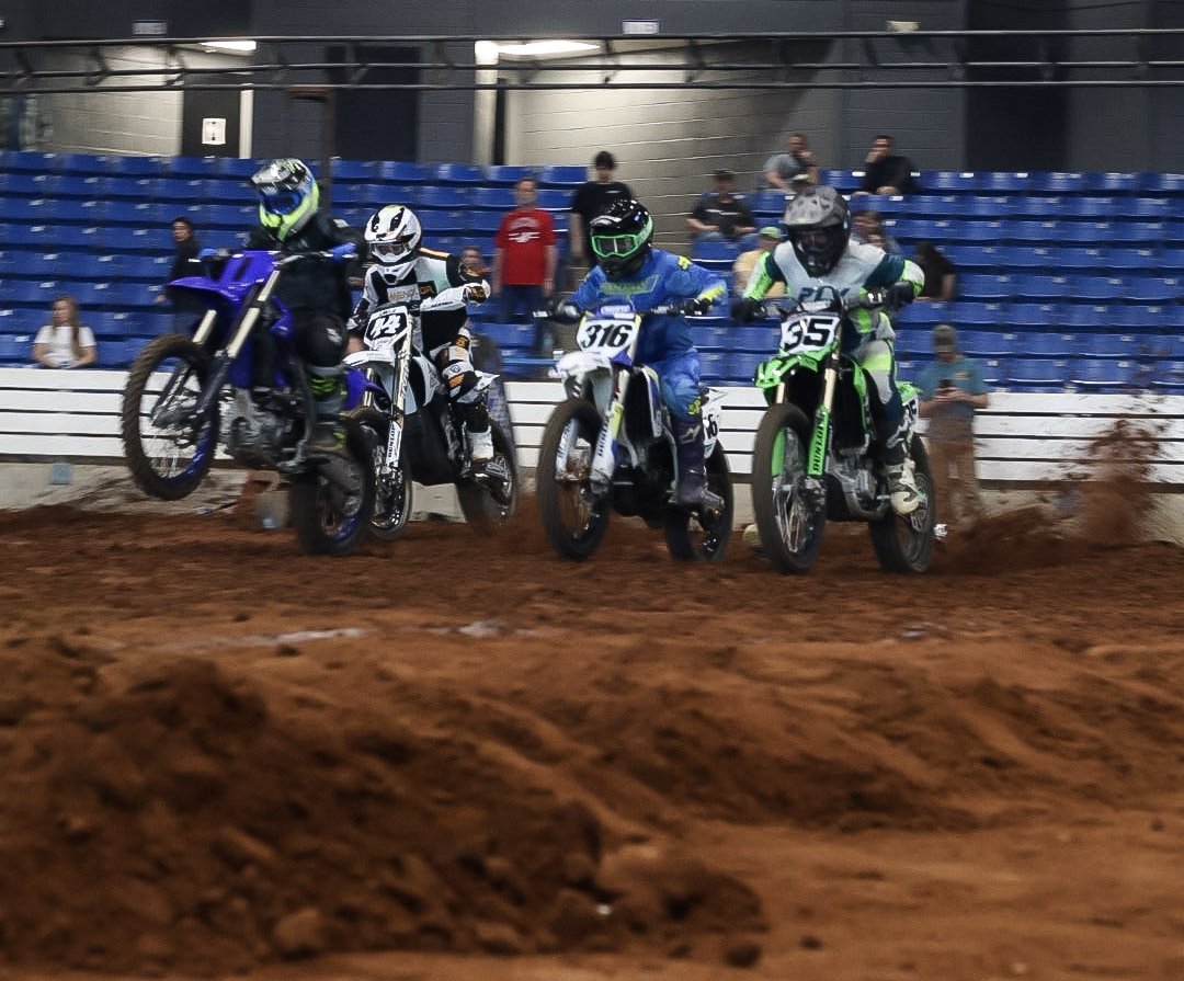 Four motocross riders in gear racing on an indoor dirt track, with one rider mid-air and the others on the ground, spectators watching in the bleachers.