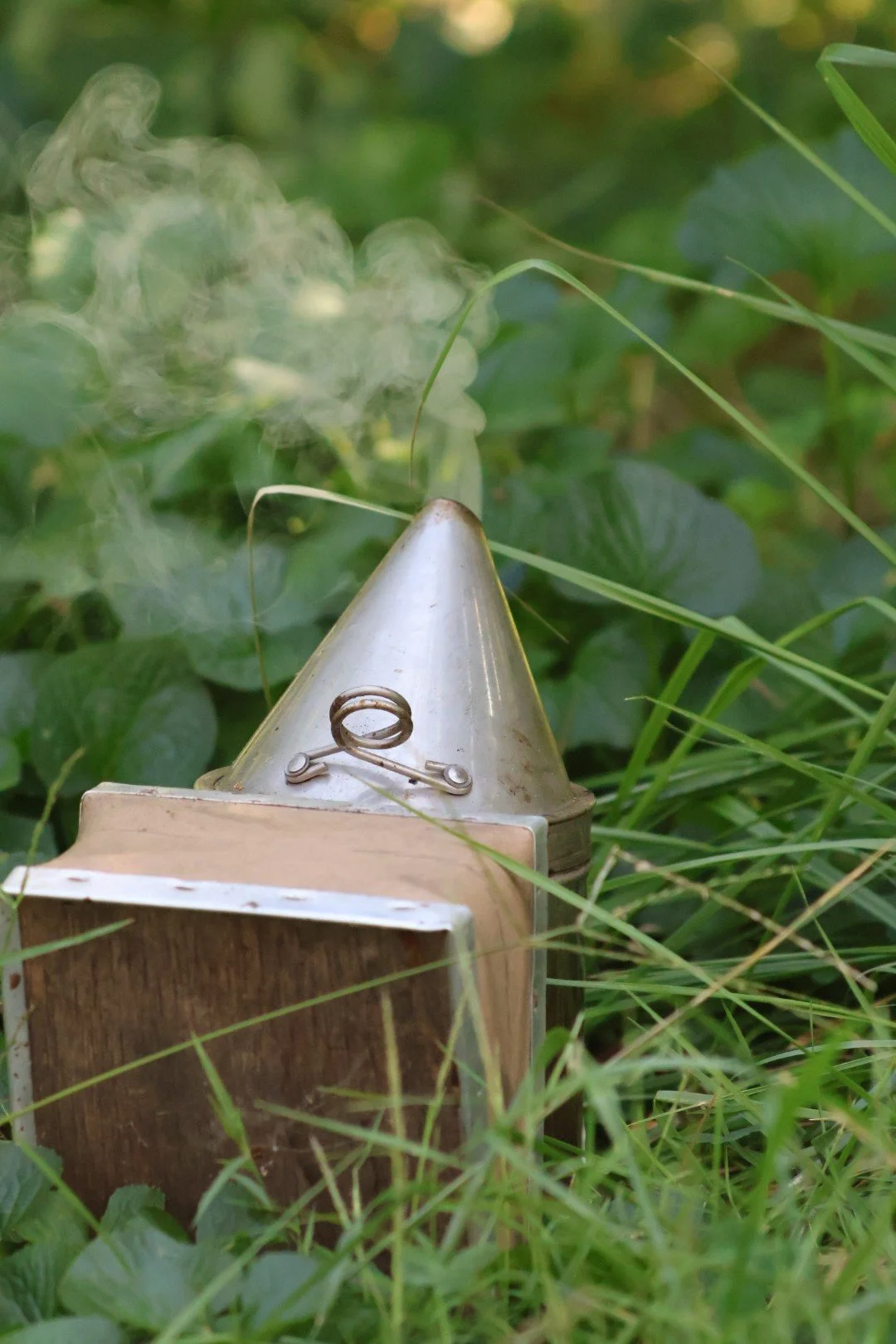 Close-up of a small metal smoke generator emitting white smoke, surrounded by green grass and foliage.