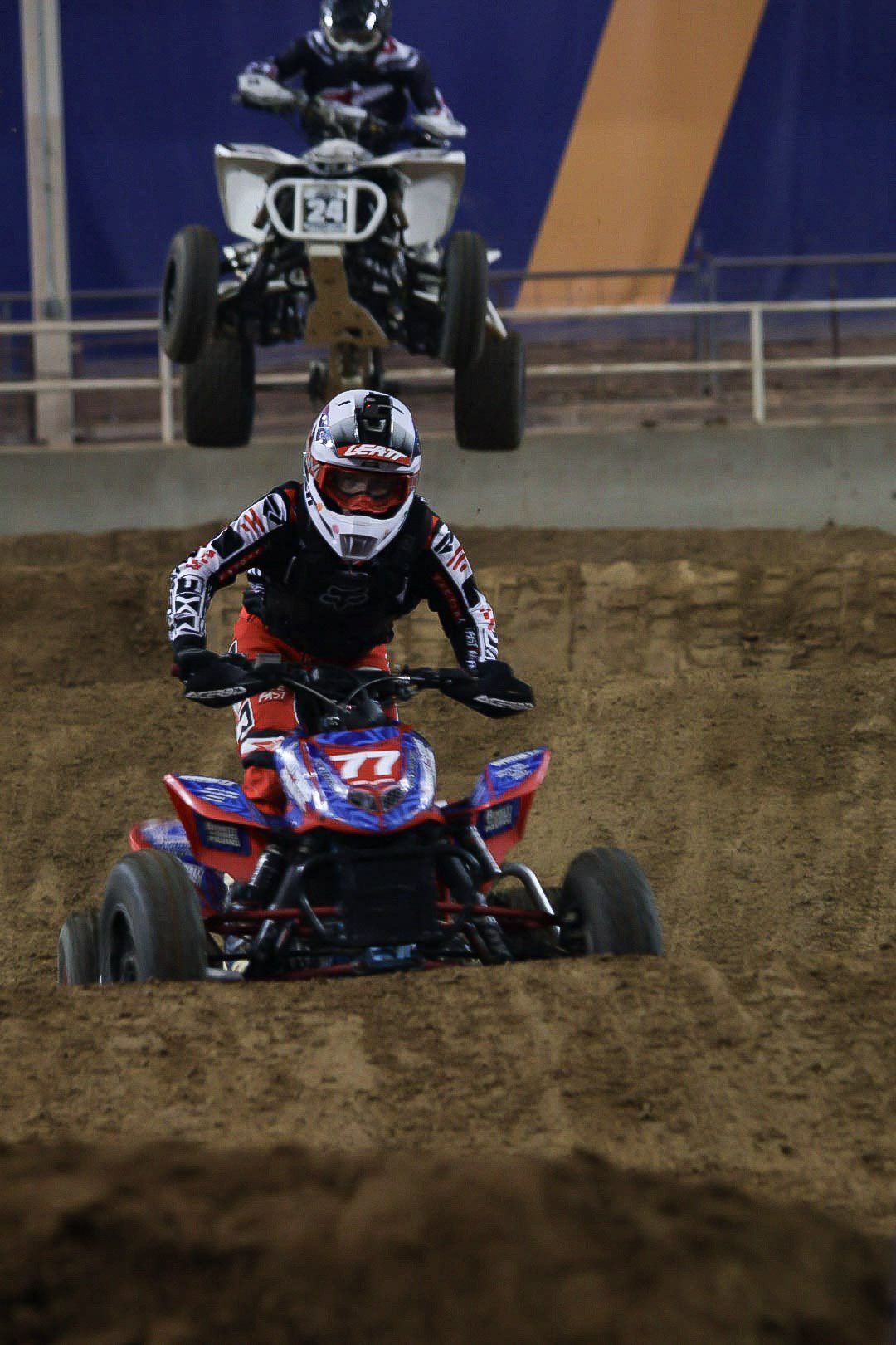 Motocross riders racing on a dirt track with one rider jumping over another in mid-air.