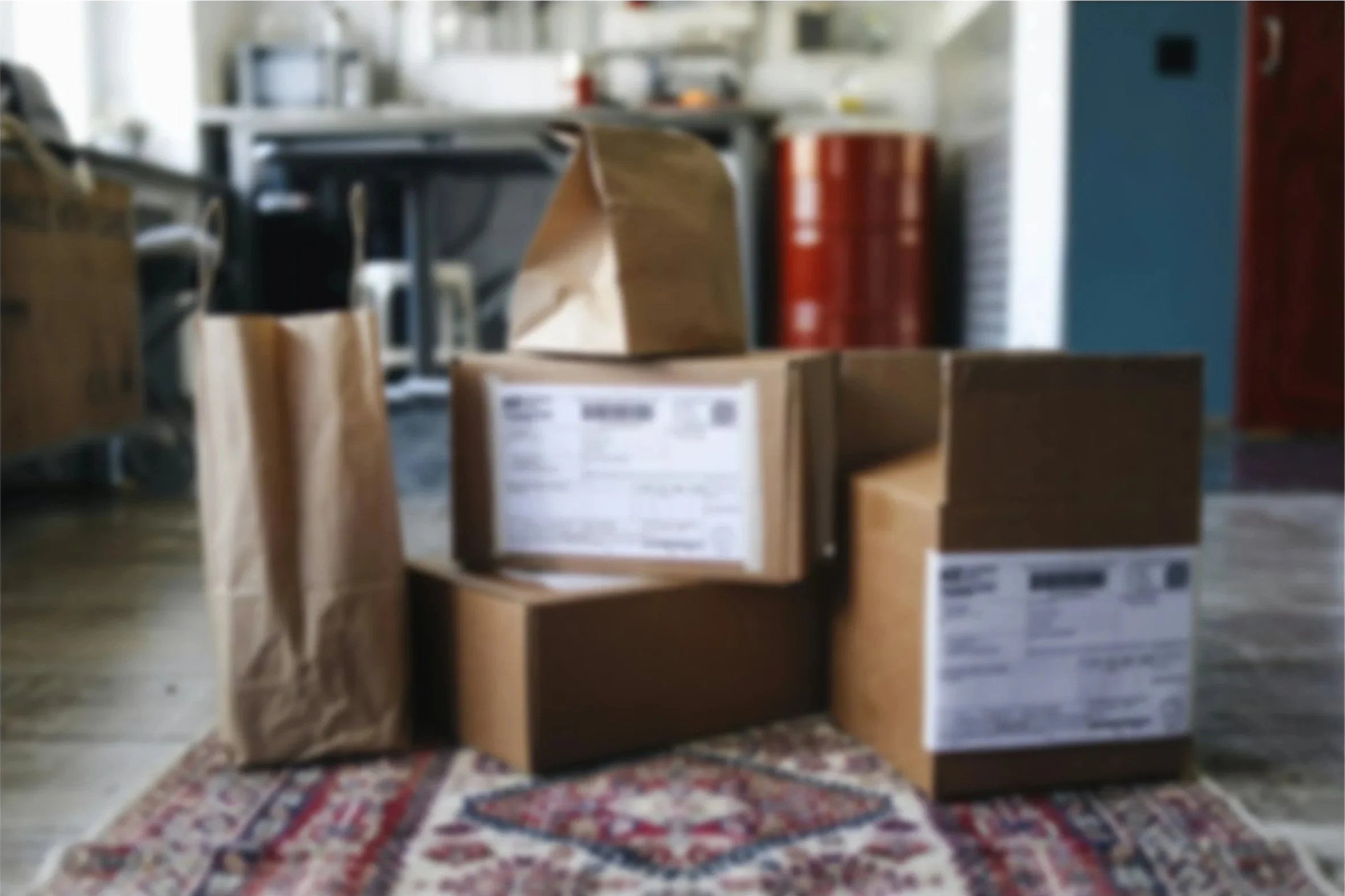 Stacked cardboard boxes and shipping packages with shipping labels on a Persian rug/textile in a room with a kitchen in the background.
