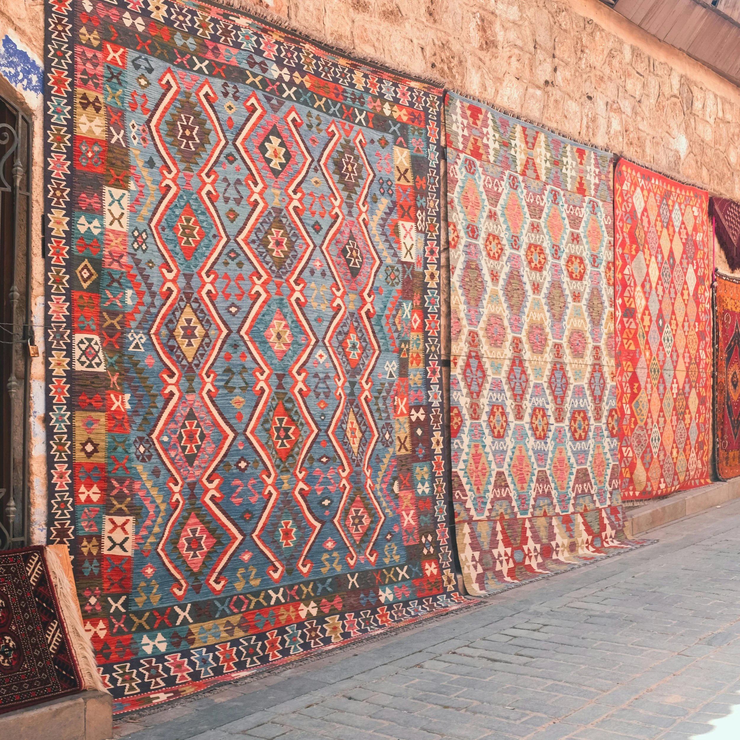 A colorful, patterned rug displayed against a brick wall on a stone floor, featuring geometric shapes and vibrant colors.