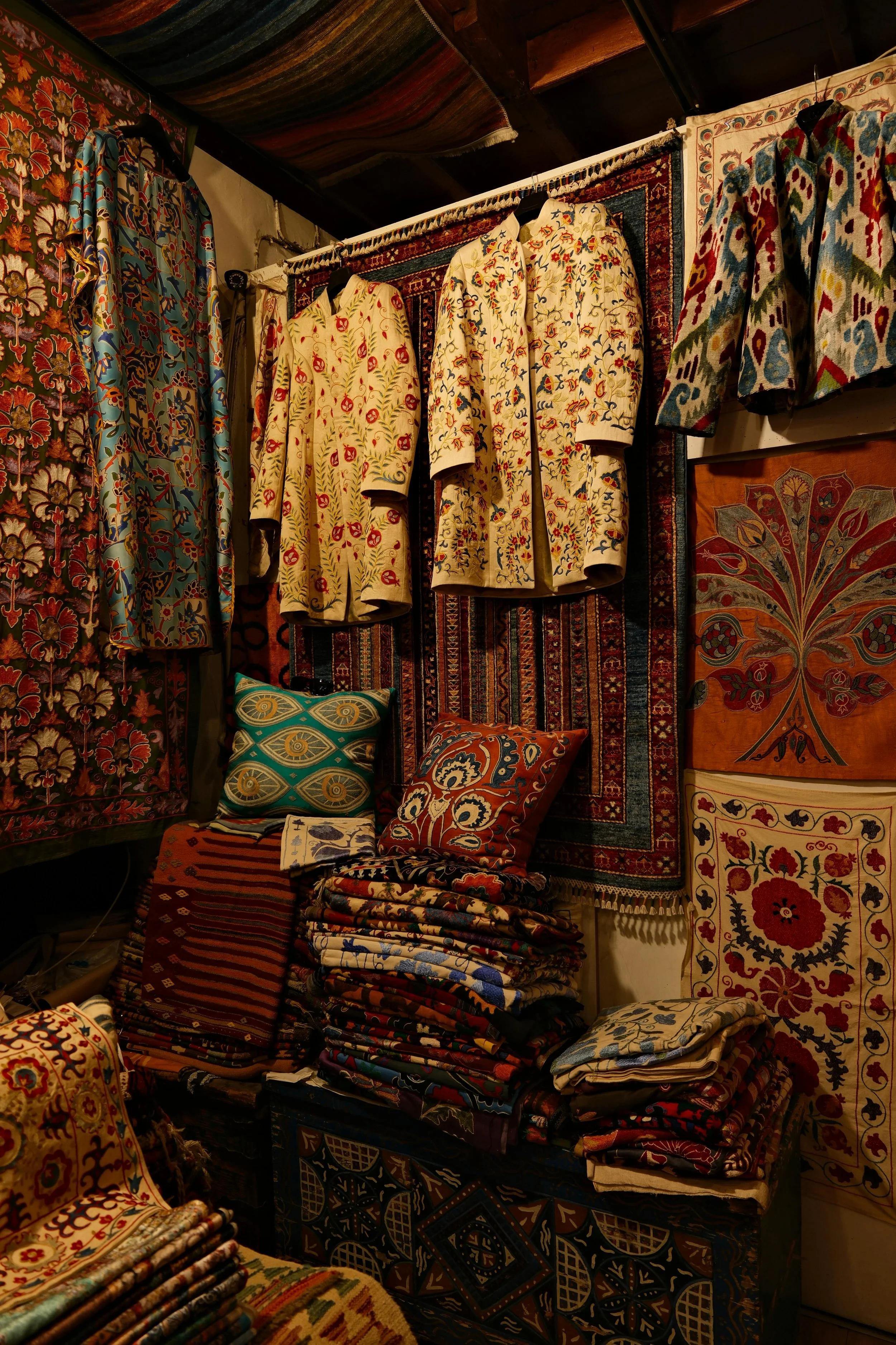 Display of various colorful patterned textiles, including embroidered and woven rugs, and fabric coats with floral and geometric designs, in a cozy indoor setting.