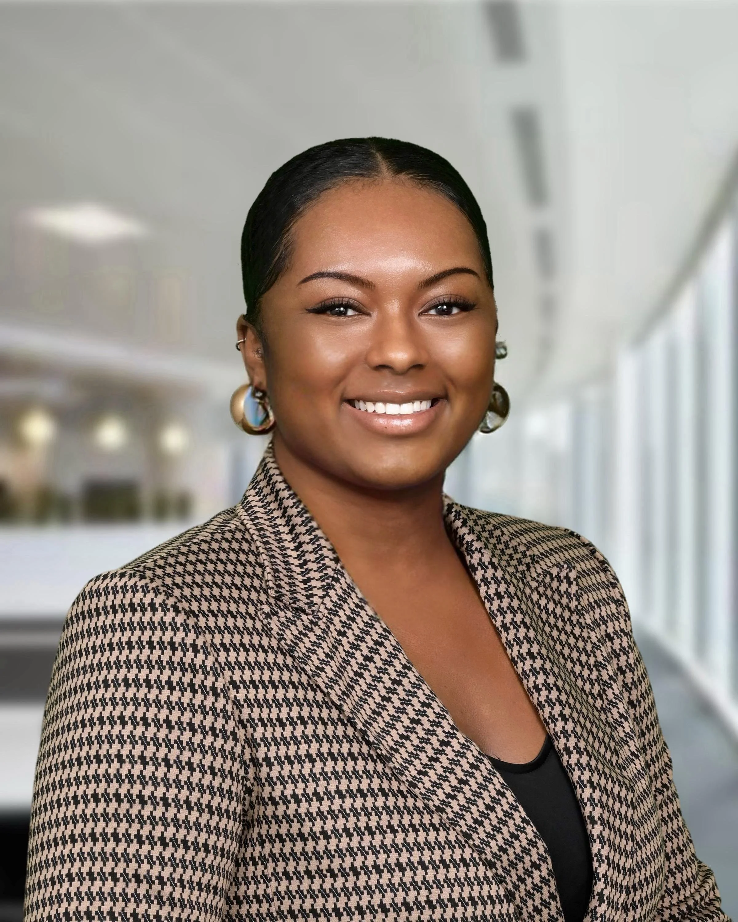 Close-up of smiling woman with dark hair, wearing earrings and a houndstooth patterned blazer, in a modern office environment.