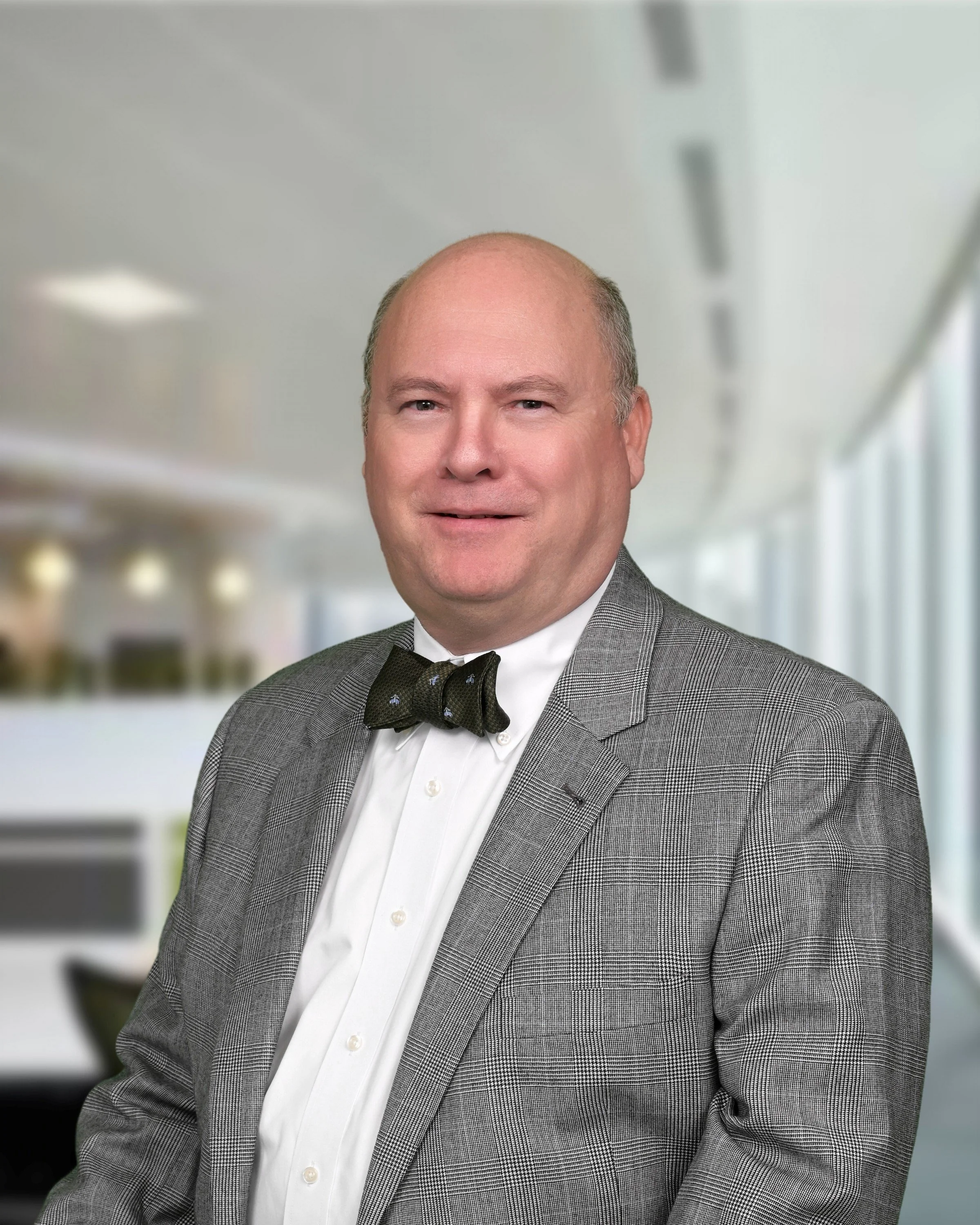 A middle-aged man dressed in a gray checkered suit, white shirt, and black bow tie smiling in an indoor setting with blurred background.