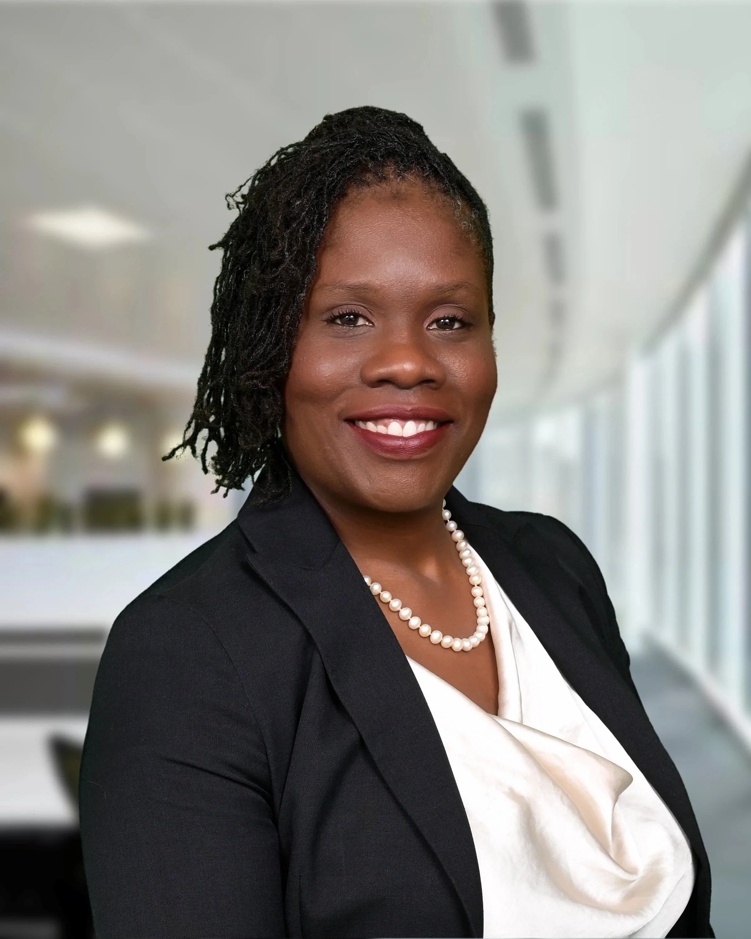 A professional woman wearing a suit and pearl necklace, smiling in an office setting with a blurred background of office lights and windows.