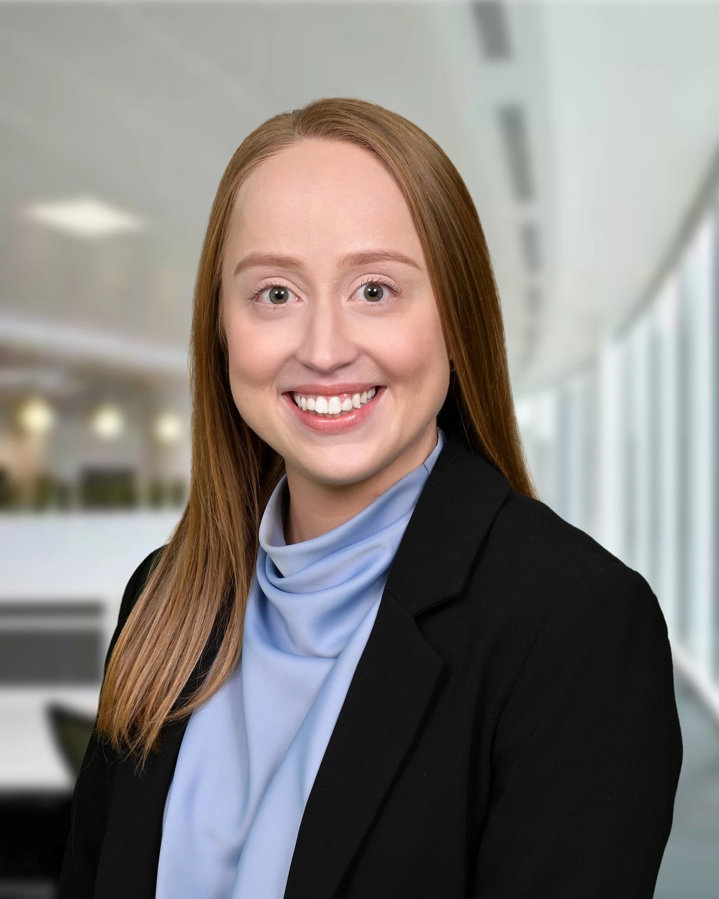 Close-up of a smiling woman with red hair, light skin, blue eyes, wearing a black blazer and a light blue scarf, in a modern office setting.