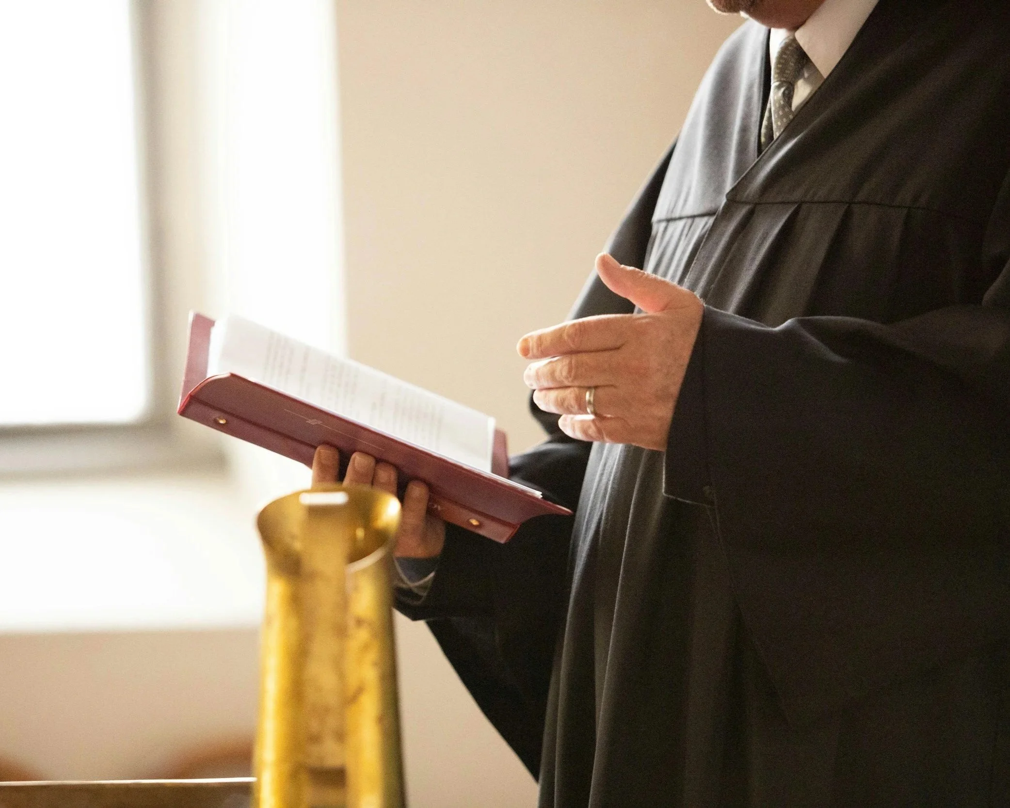 A man in a black suit and tie holding a red book or folder, with a gold-colored post or pole in the foreground, standing indoors near a window.