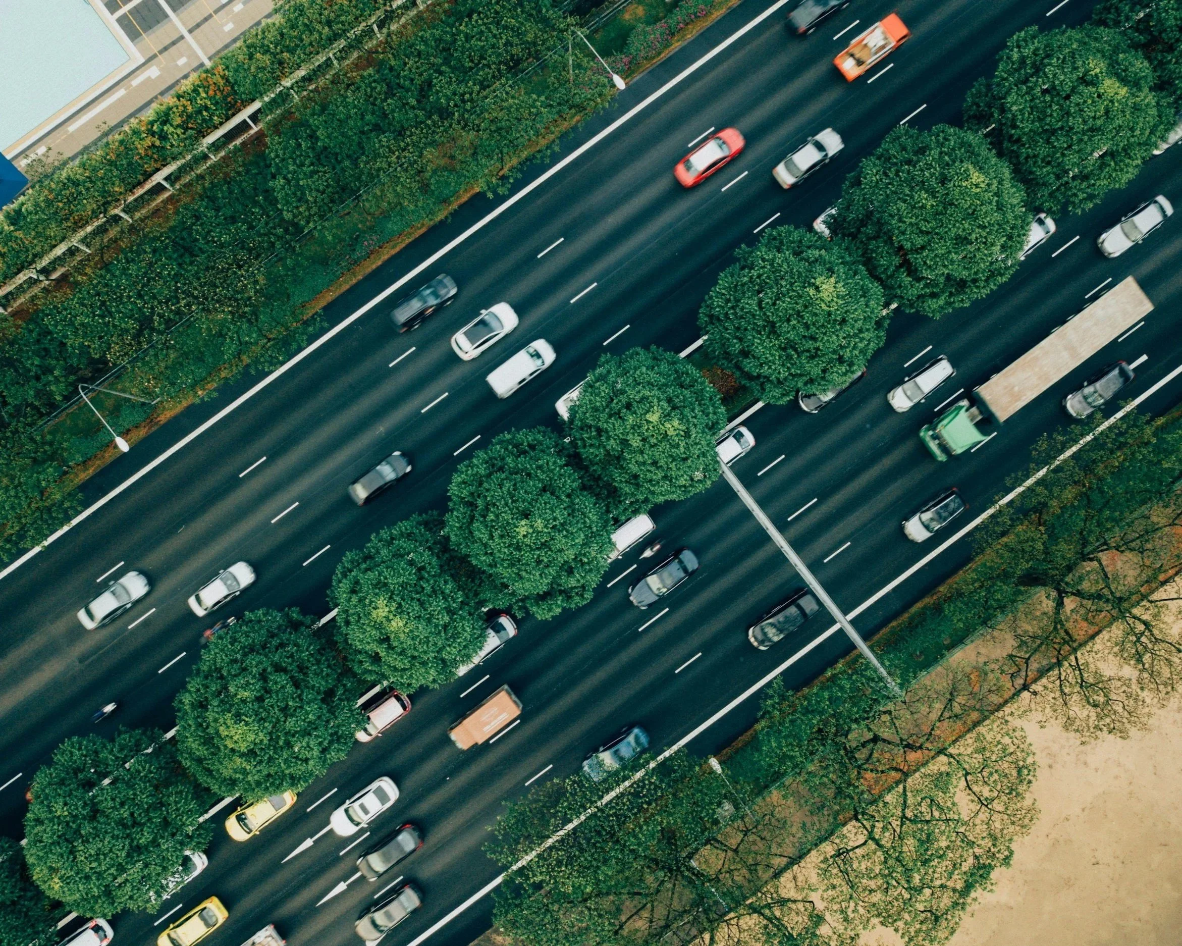 An aerial view of a multi-lane city street with cars traveling in both directions, lined with green trees and streetlights.
