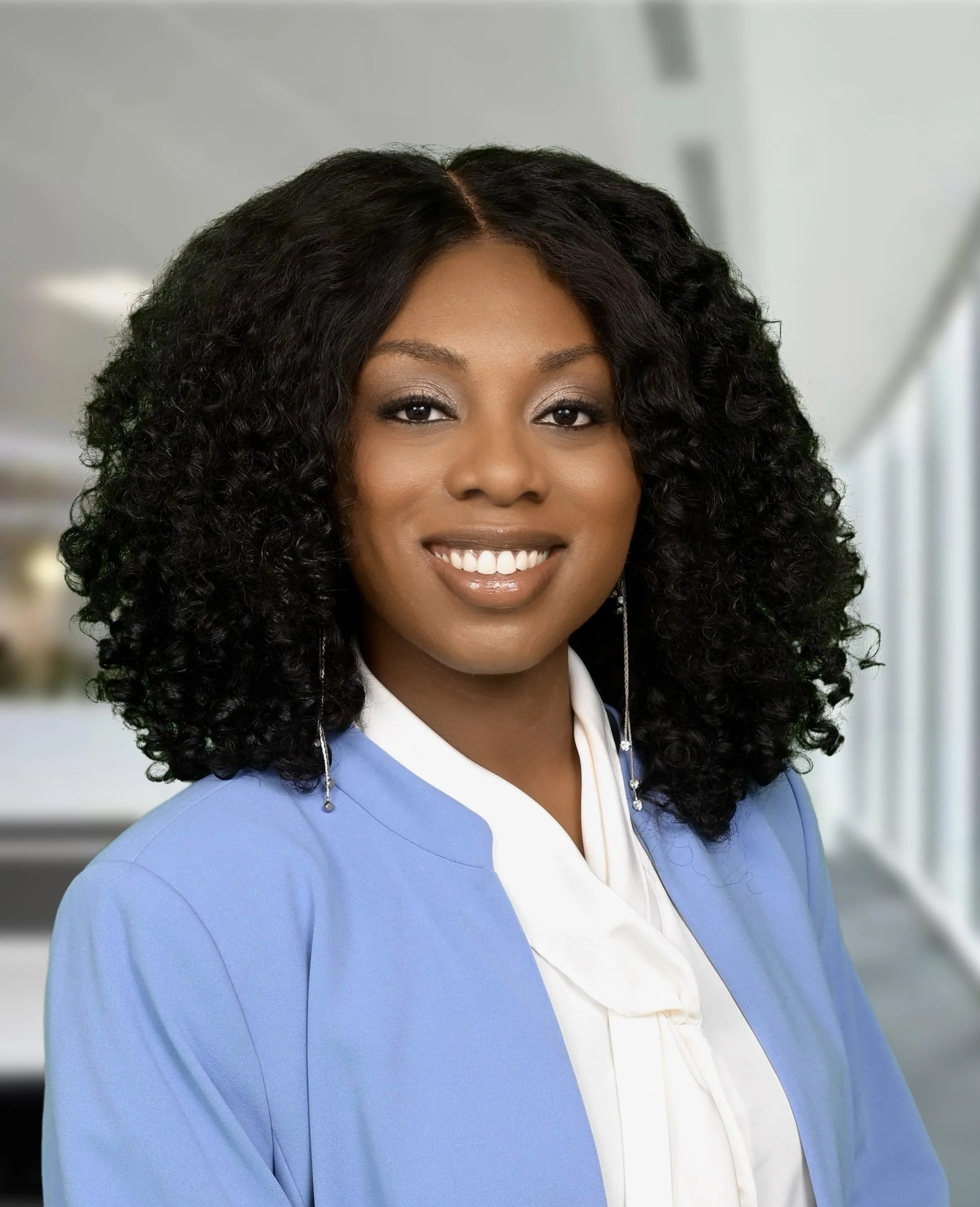 Close-up of a woman with curly black hair, smiling, wearing a blue blazer over a white blouse, in an indoor setting.