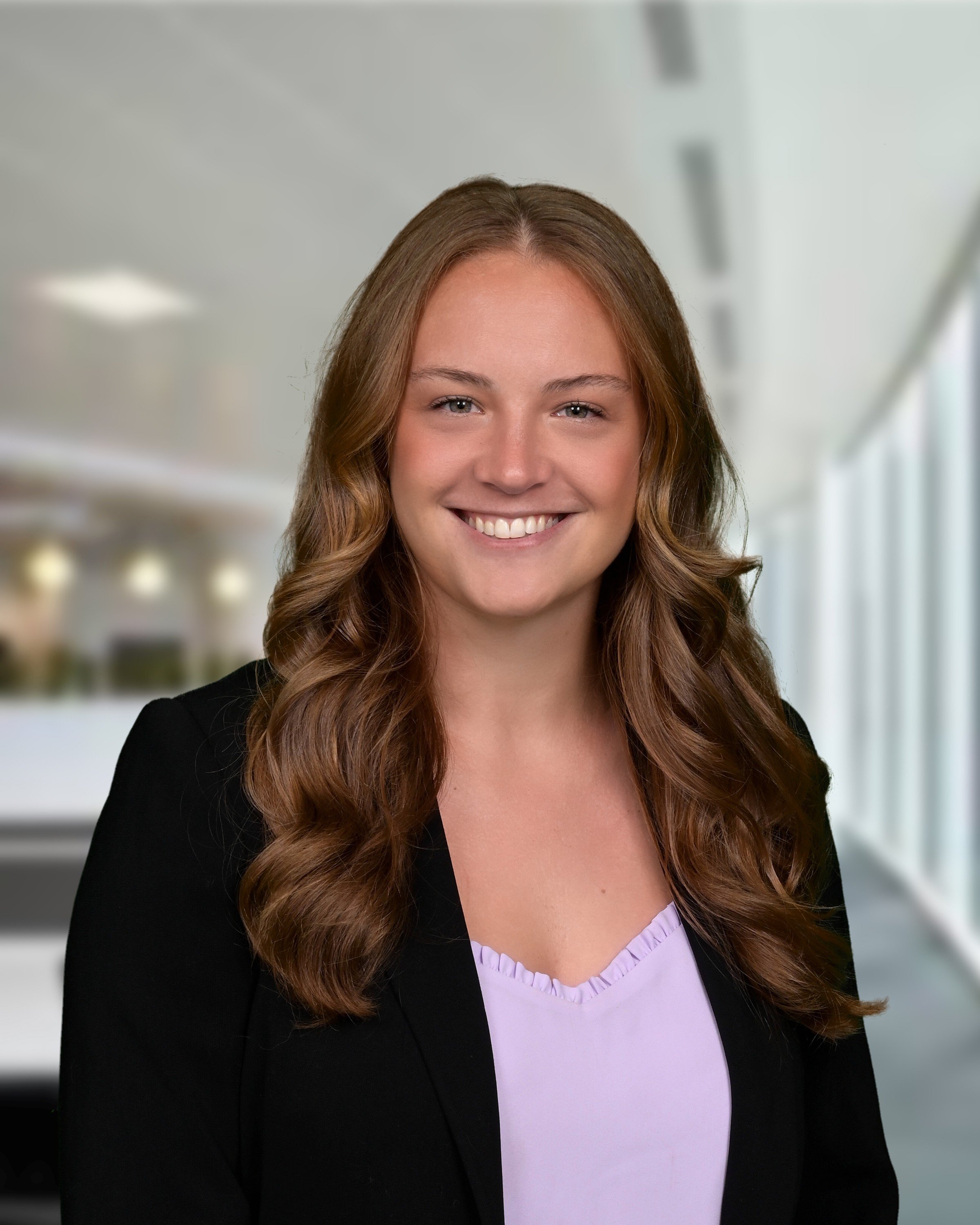 Young woman with long, wavy brown hair smiling, wearing a black blazer over a lavender top, standing in a brightly lit office corridor.