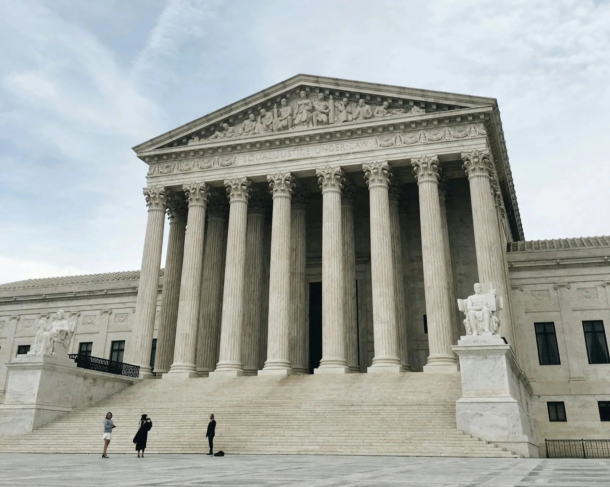 The United States Supreme Court building with large steps, tall columns, and sculptural friezes at the top. Several people are standing and walking on the steps.