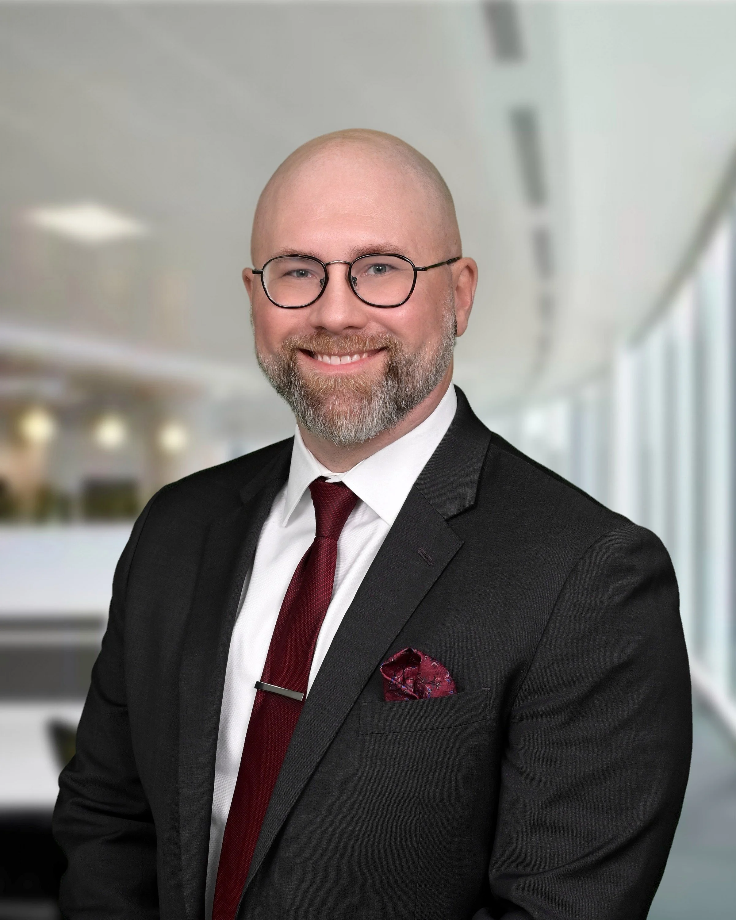 A professional man with a beard, glasses, dressed in a black suit, white shirt, and burgundy tie, smiling for a portrait against a blurred indoor background.