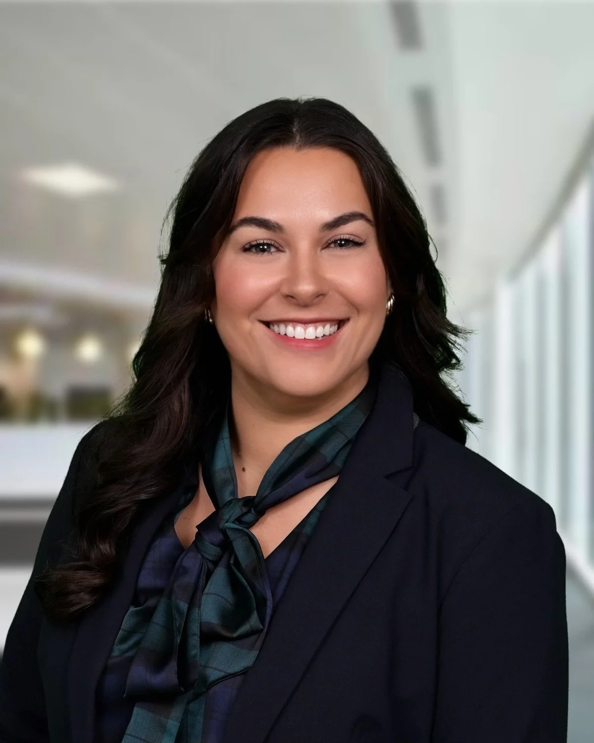 Professional woman with dark hair, wearing a dark blazer and a blue plaid scarf, smiling in an office setting.