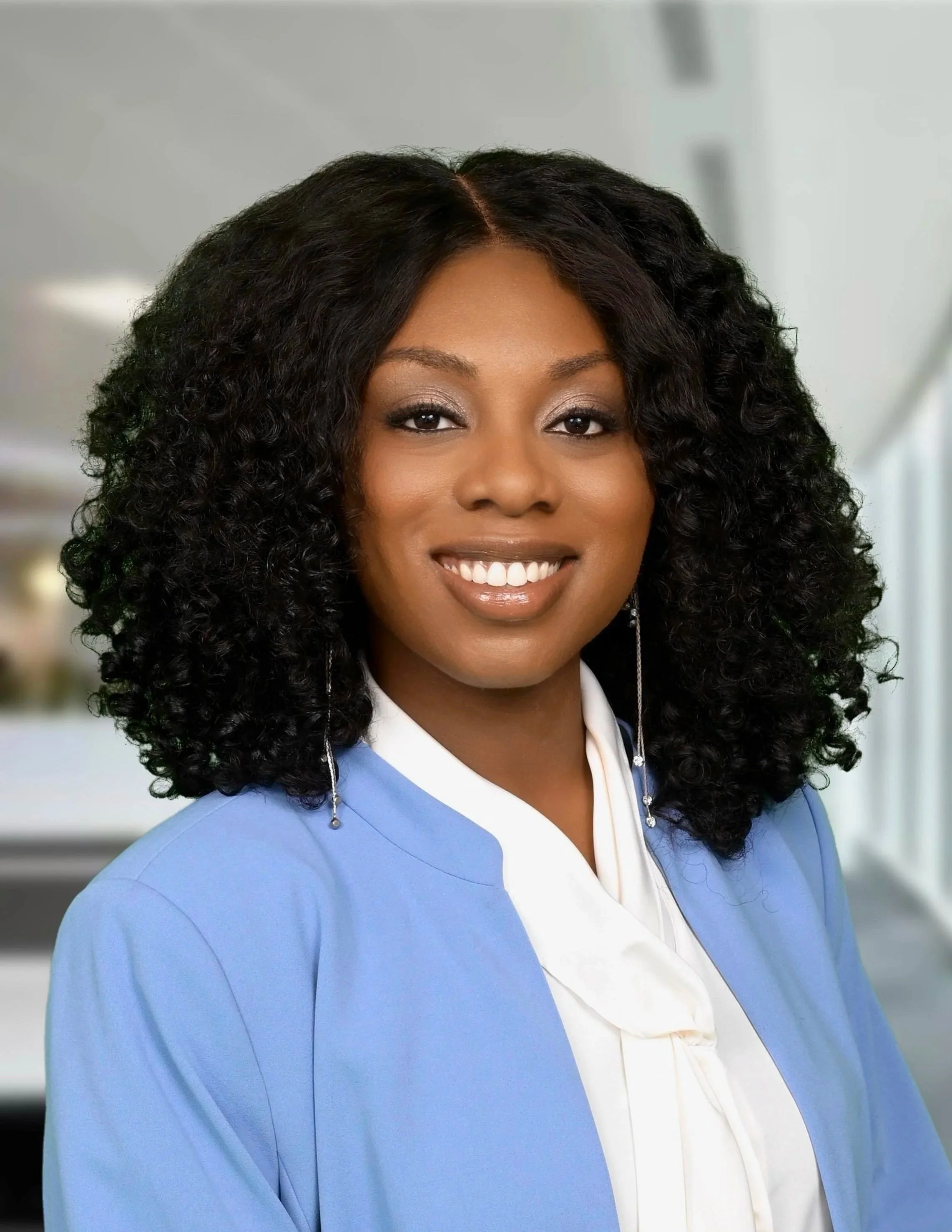 A woman with dark curly hair smiling, wearing a blue blazer and white blouse, in an indoor setting.