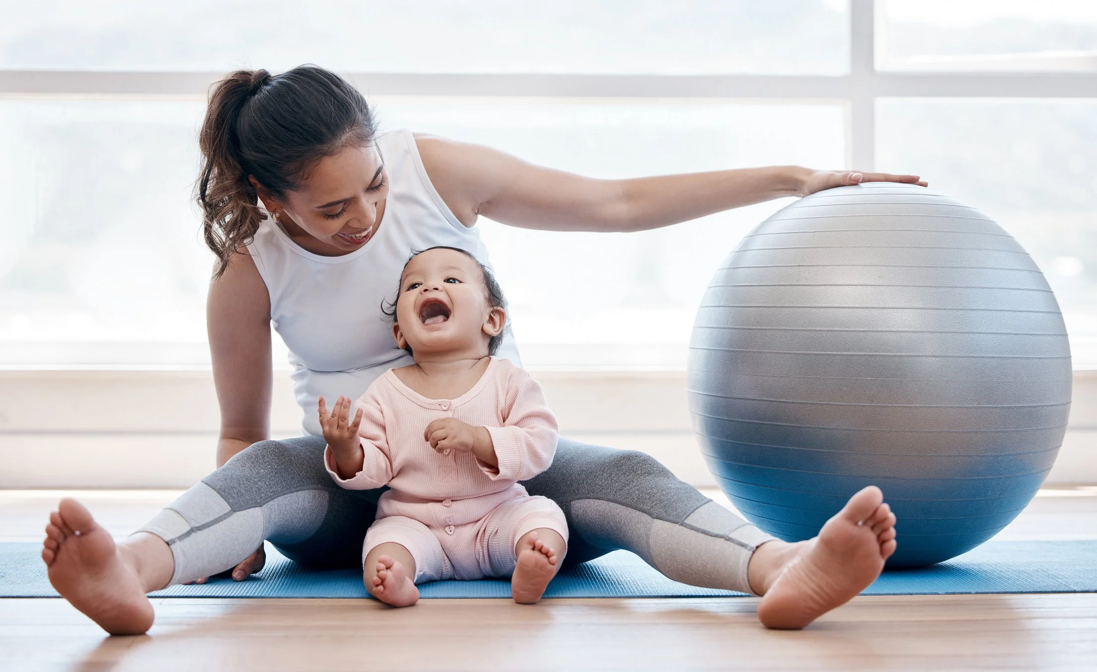 Woman and child engaging in yoga or playful activity on a blue mat in a bright room with large windows, with a large exercise ball nearby.