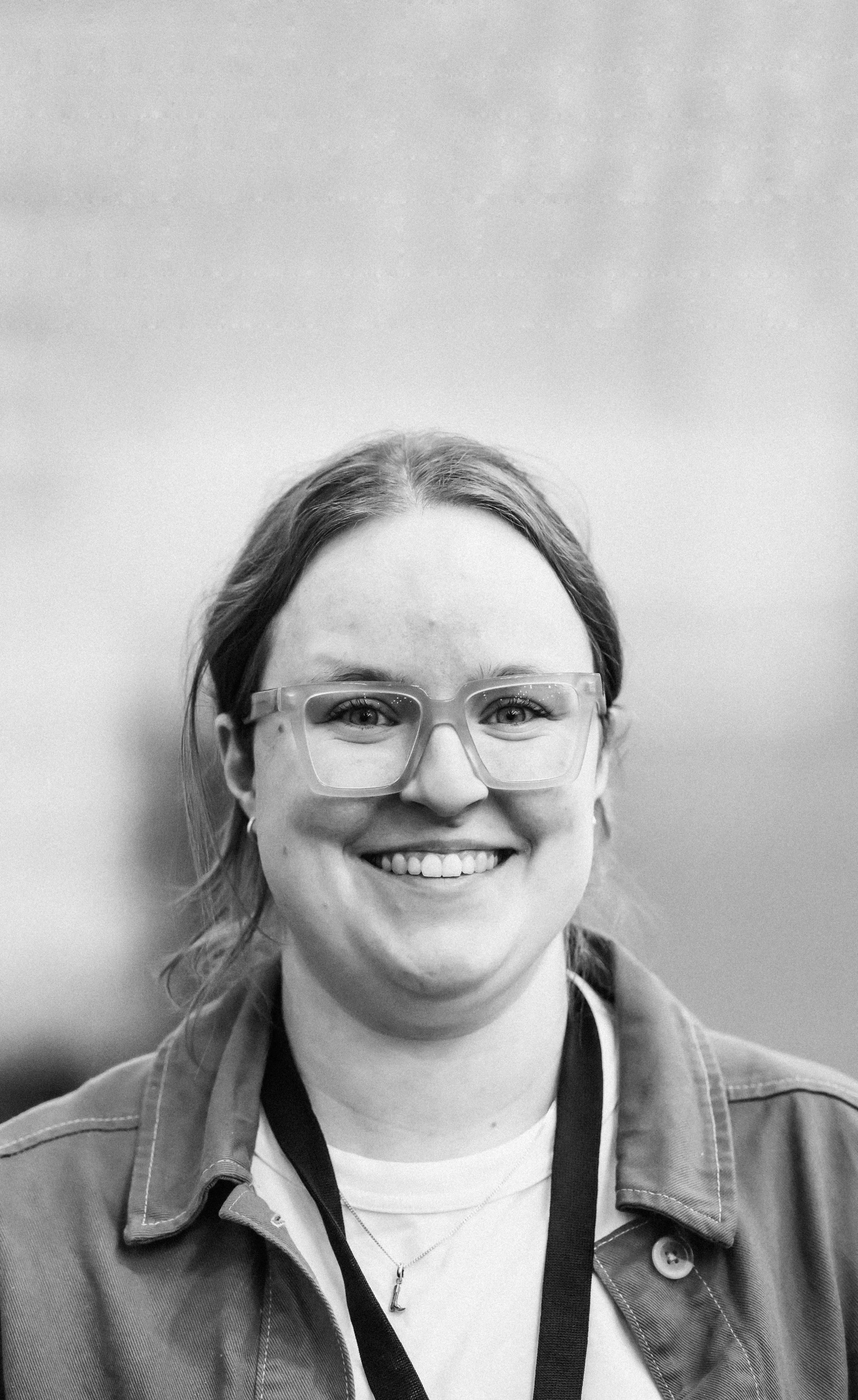 Portrait en noir et blanc d'une jeune femme souriante portant des lunettes et une veste, avec un fond neutre.