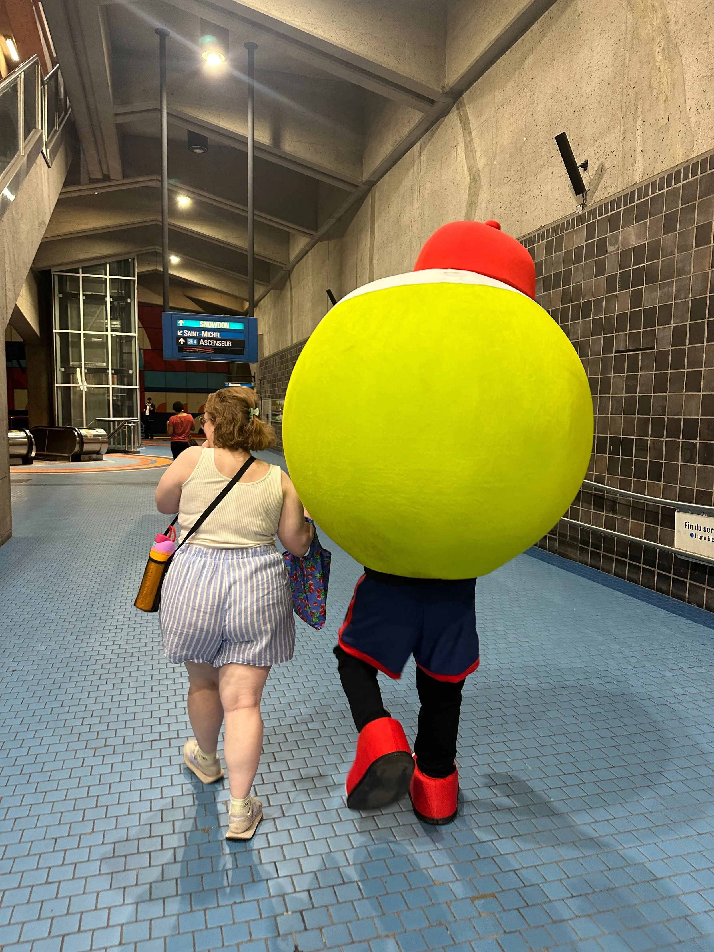 Une femme marche dans une station de métro avec un homme déguisé en super-héros portant un costume avec une grosse boule verte sur le dos. La station a un sol en carreaux bleus et des panneaux d'indication.