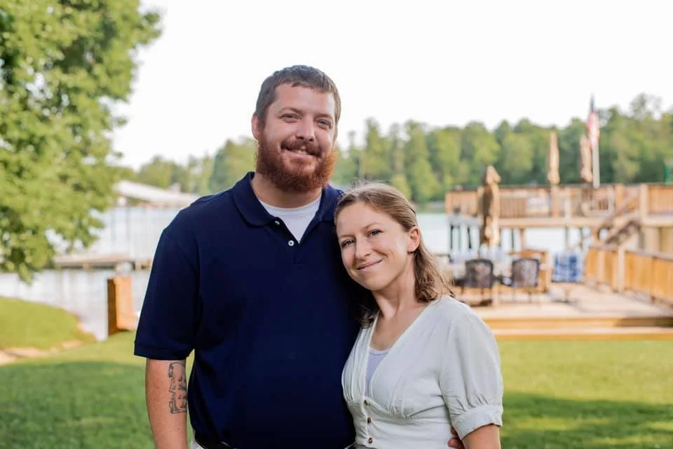 A smiling man with a beard and a woman with shoulder-length hair standing outdoors near a lake or river with a wooden deck and green trees in the background.