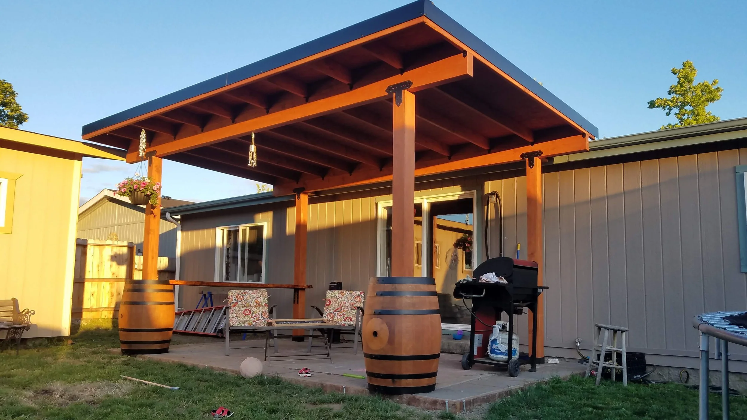 A gazebo attached to a house offers cover for lawn chairs, a decorate barrel, and a BBQ