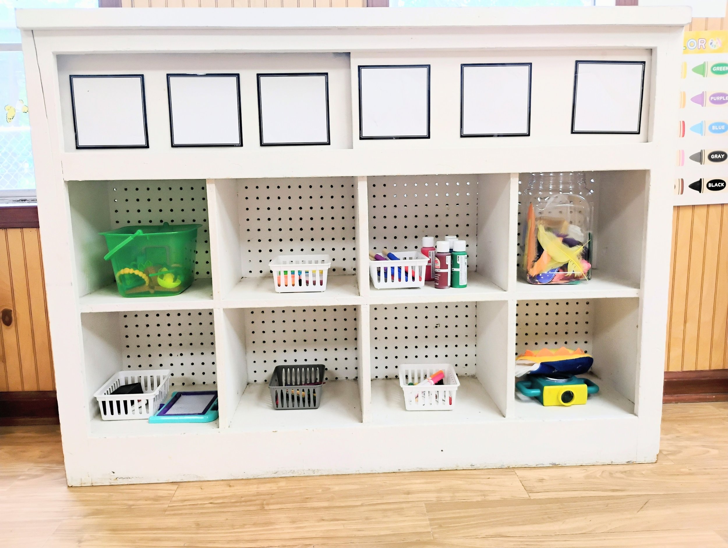 Empty white wooden cubby shelf with small plastic baskets, bottles of glue, and craft supplies, situated on a wooden floor with wooden paneling on the walls.
