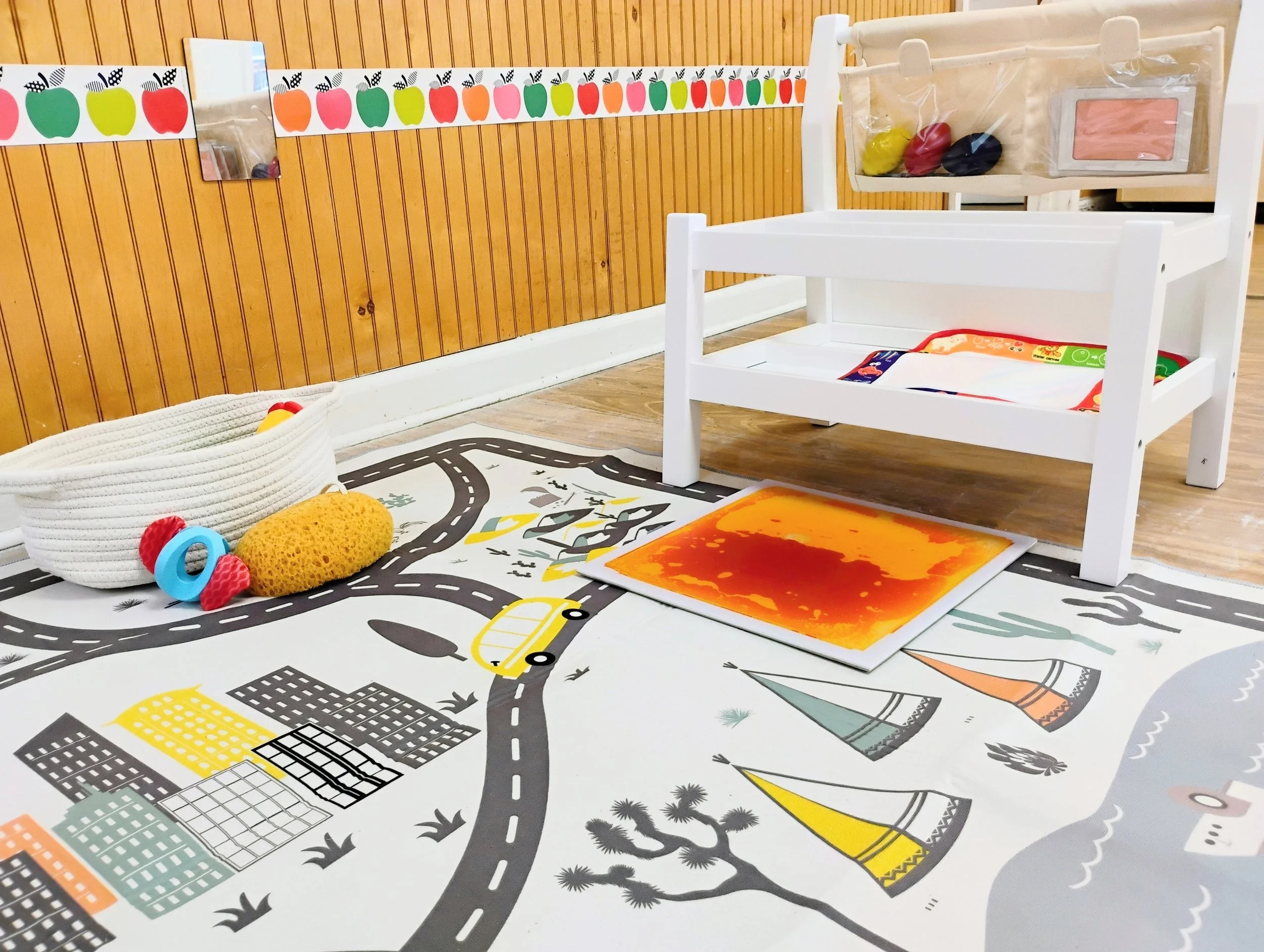 Colorful decor and toys in a children's play area, including a white wooden bed, a woven basket with balls, a city-themed rug, and wall decorations of apples and a mirror.