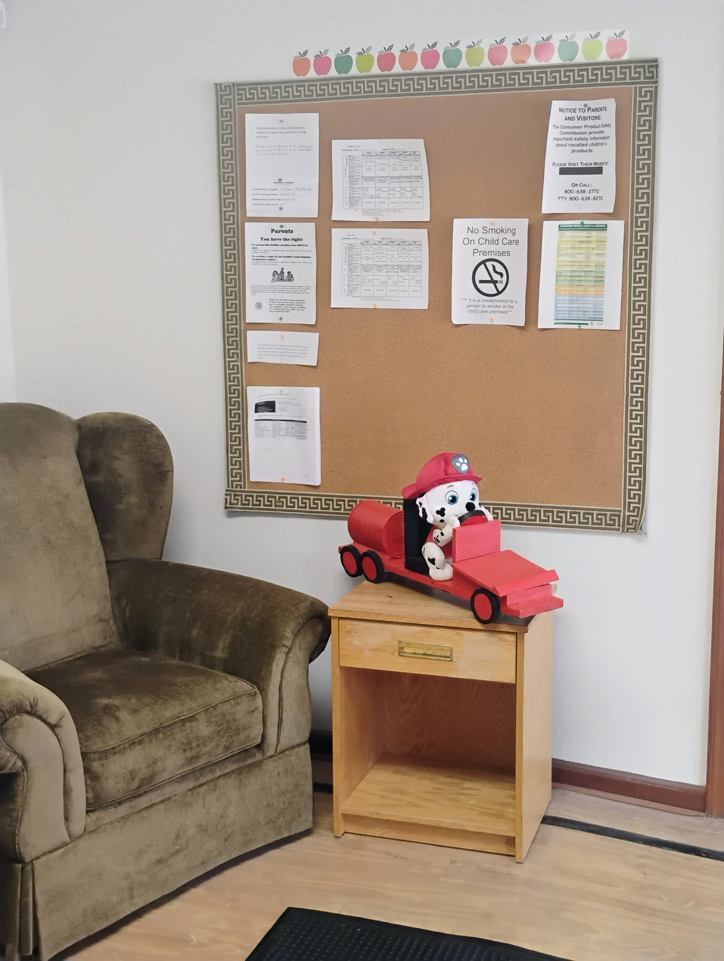 A cork bulletin board with various notices, charts, and notices, decorated with apple and rainbow-colored paper top border. Next to the board, there is a red toy dog on a small wooden table and a brown upholstered armchair.