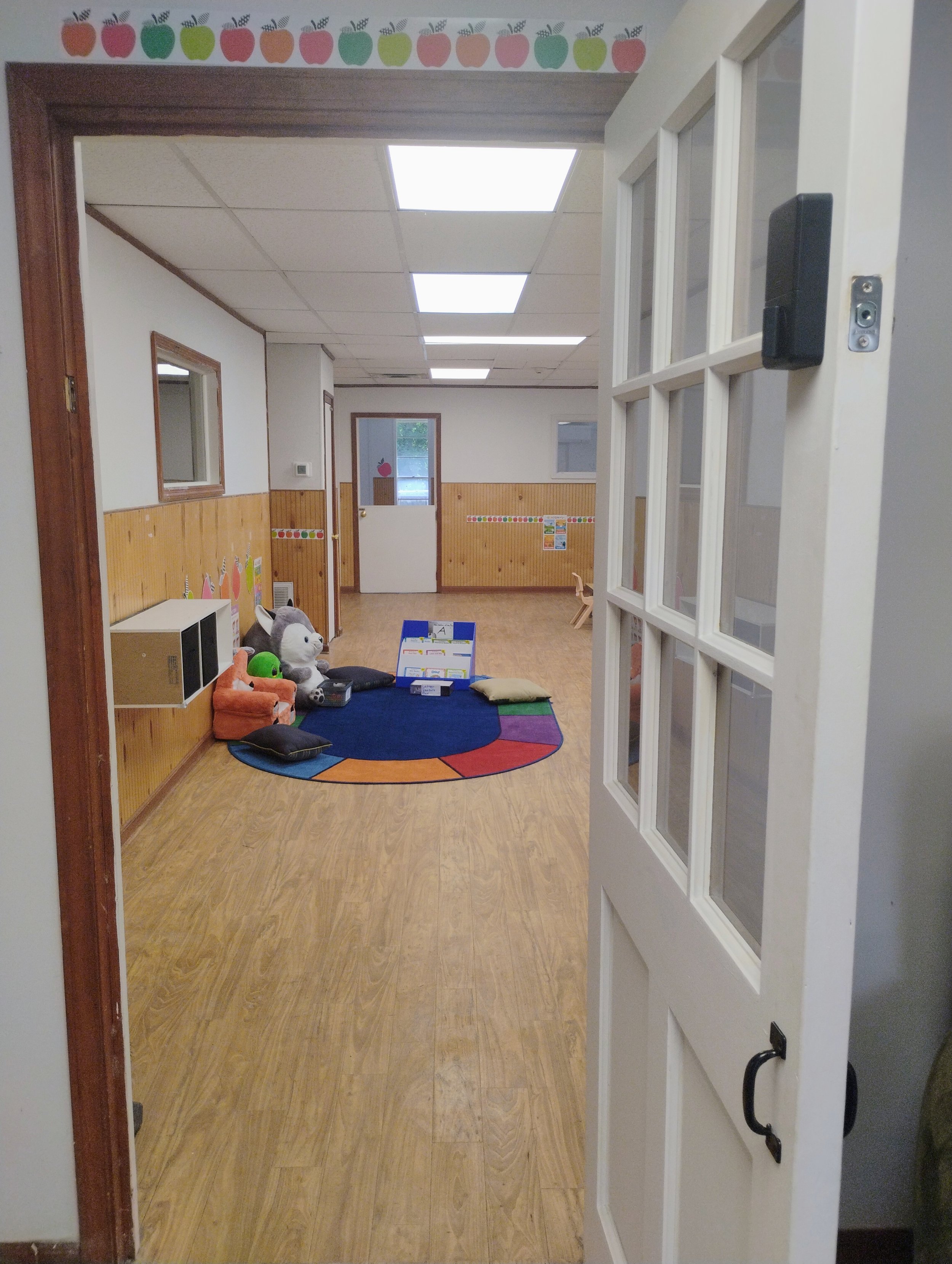 A view from a door into a room with wooden flooring, a colorful striped rug, plush toys including a panda and green character, small whiteboard or display, and walls decorated with colored apple and fruit border stickers. The ceiling has white tiles 
