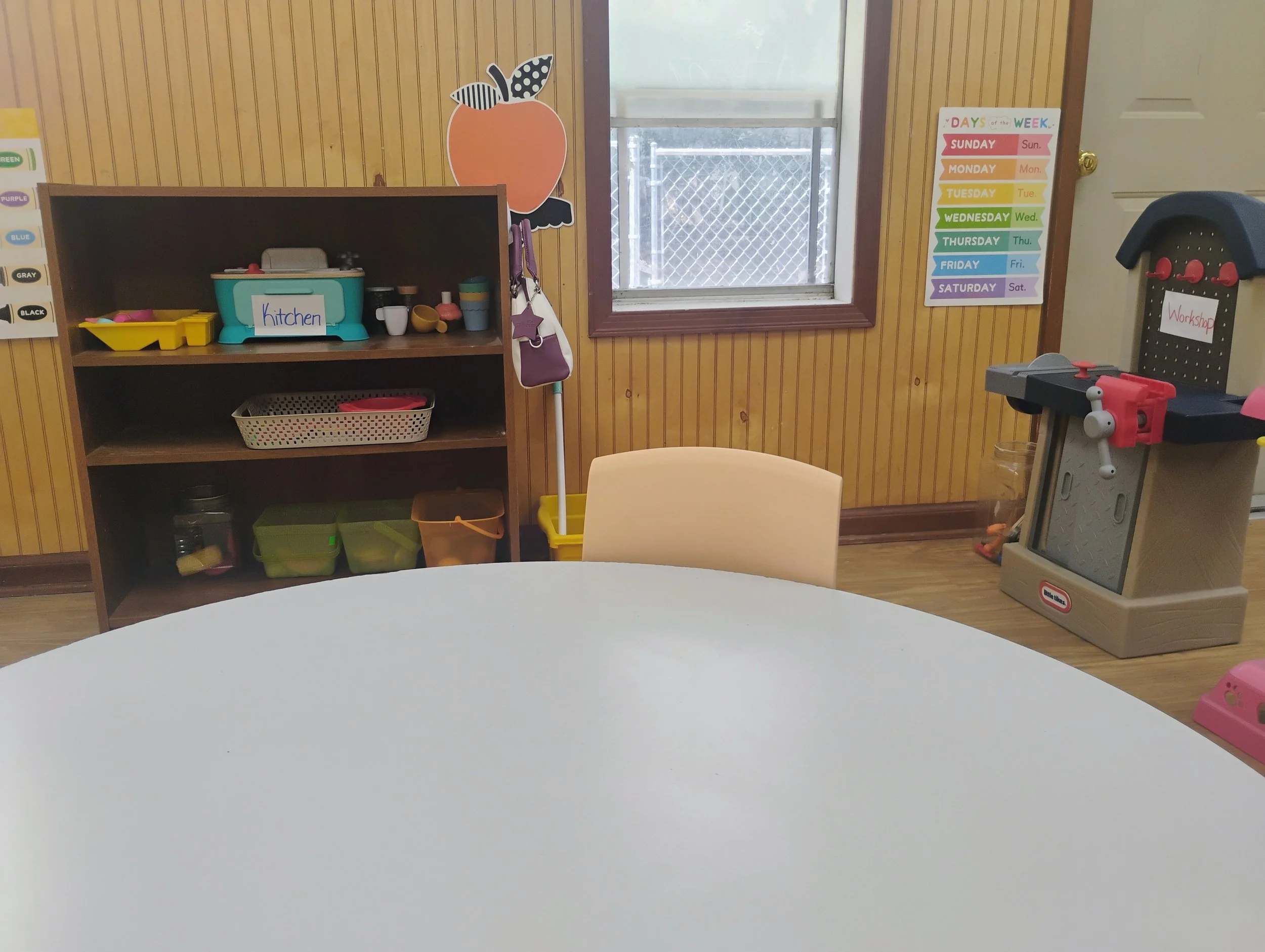 Classroom corner with wooden wall, window, colorful schedule chart, and a toy workshop set. The set includes a toy saw table and storage bins.