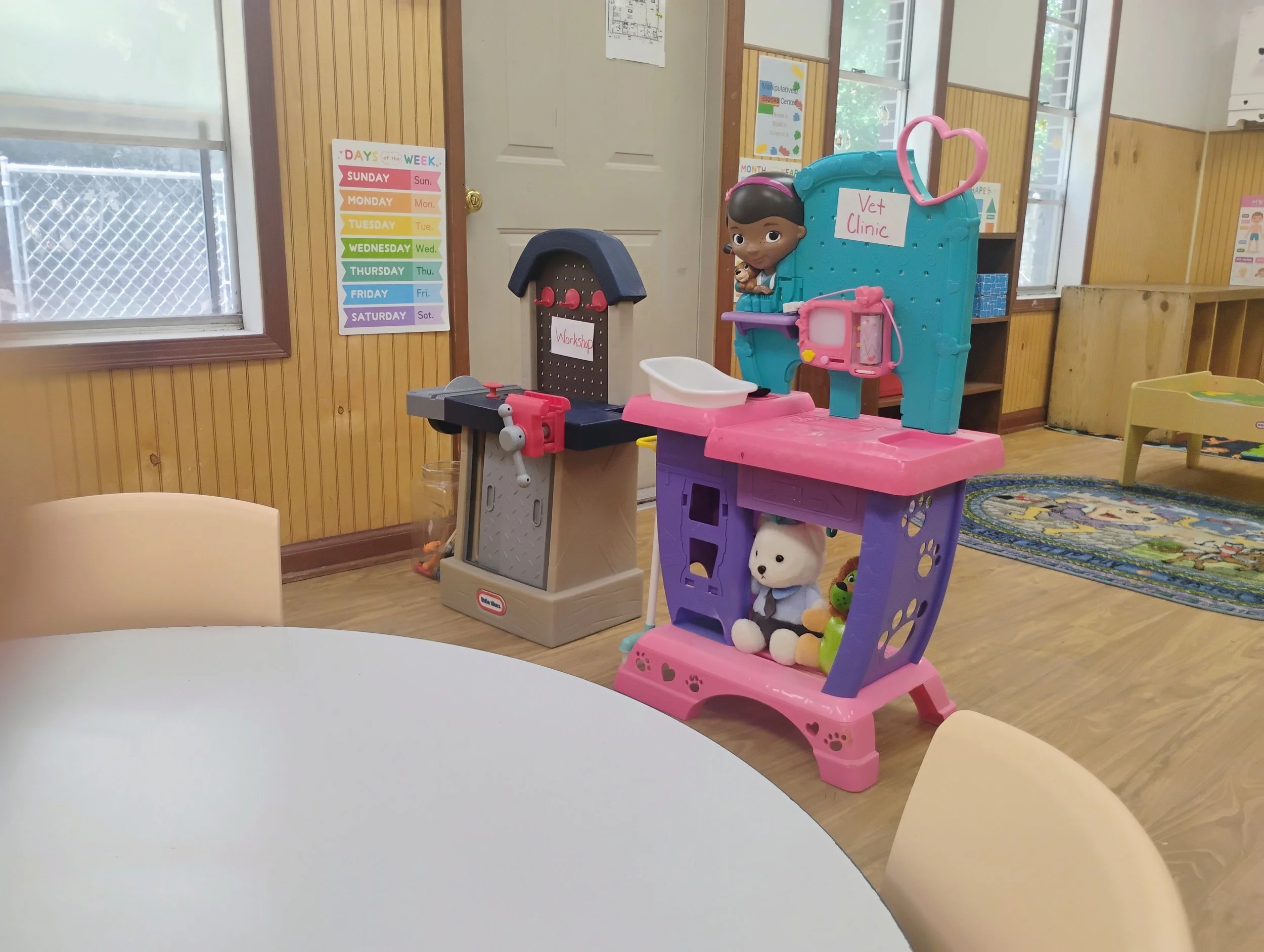 Children's play area with a pink toy shopping cart, a toy kitchen, and a toy veterinarian clinic. There are colorful charts on the wall, including a weekly days chart and a monthly calendar. The room has wooden paneling and windows letting in natural