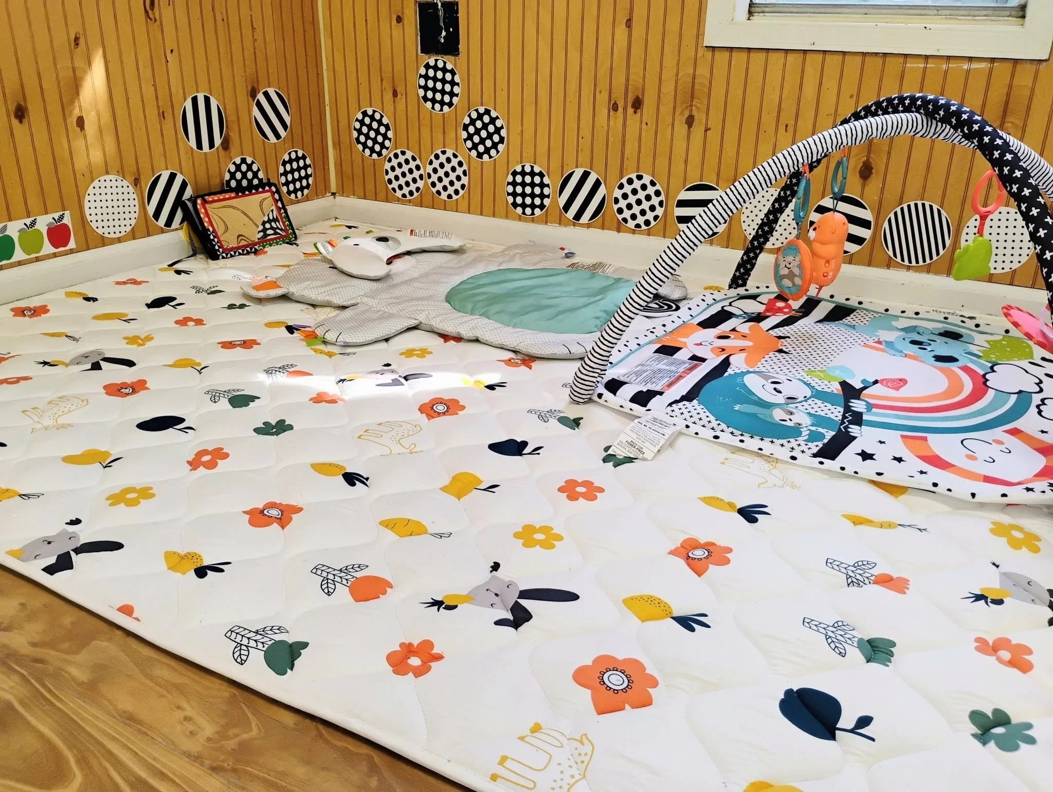 A nursery bed covered with a floral-patterned mattress, decorated with black and orange flowers. There is a green pillow, pillows with animal prints, and a colorful baby blanket. The wall behind has black and white polka dot and striped circular decorations, with a small window above.