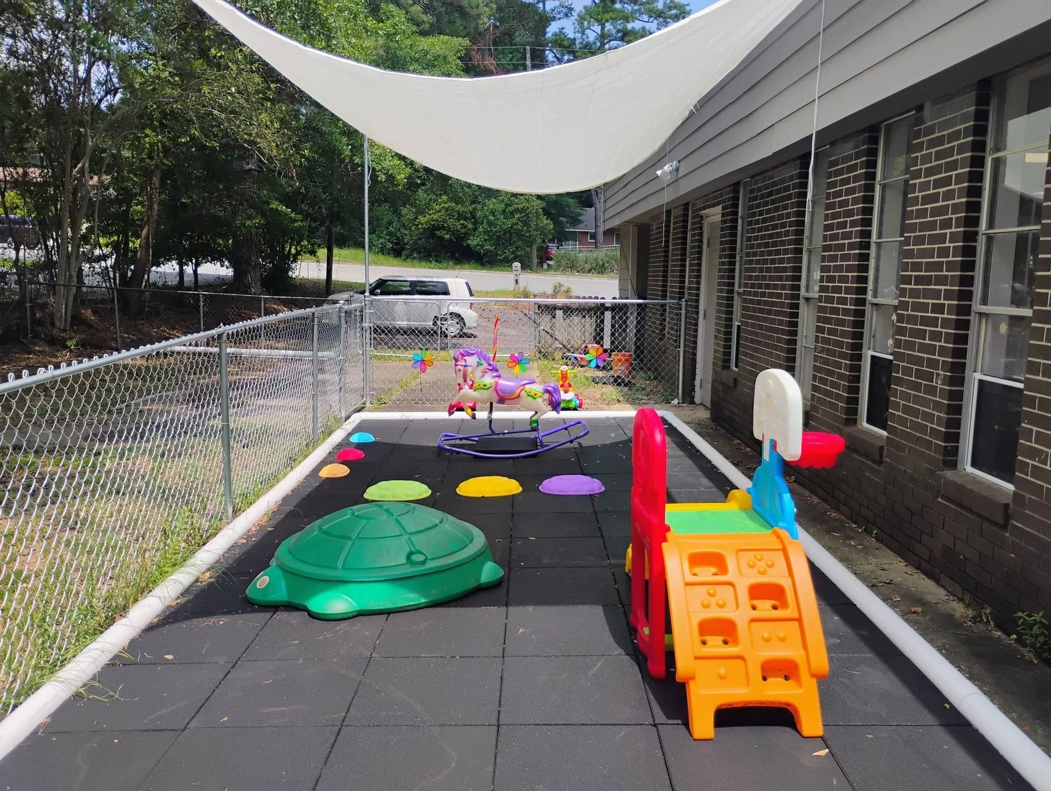 A backyard play area with a black rubber mat floor, enclosed by a white chain-link fence, containing colorful children's toys including a pink and white rocking unicorn, a red and yellow slide, a green plastic tunnel, and several circular stepping st