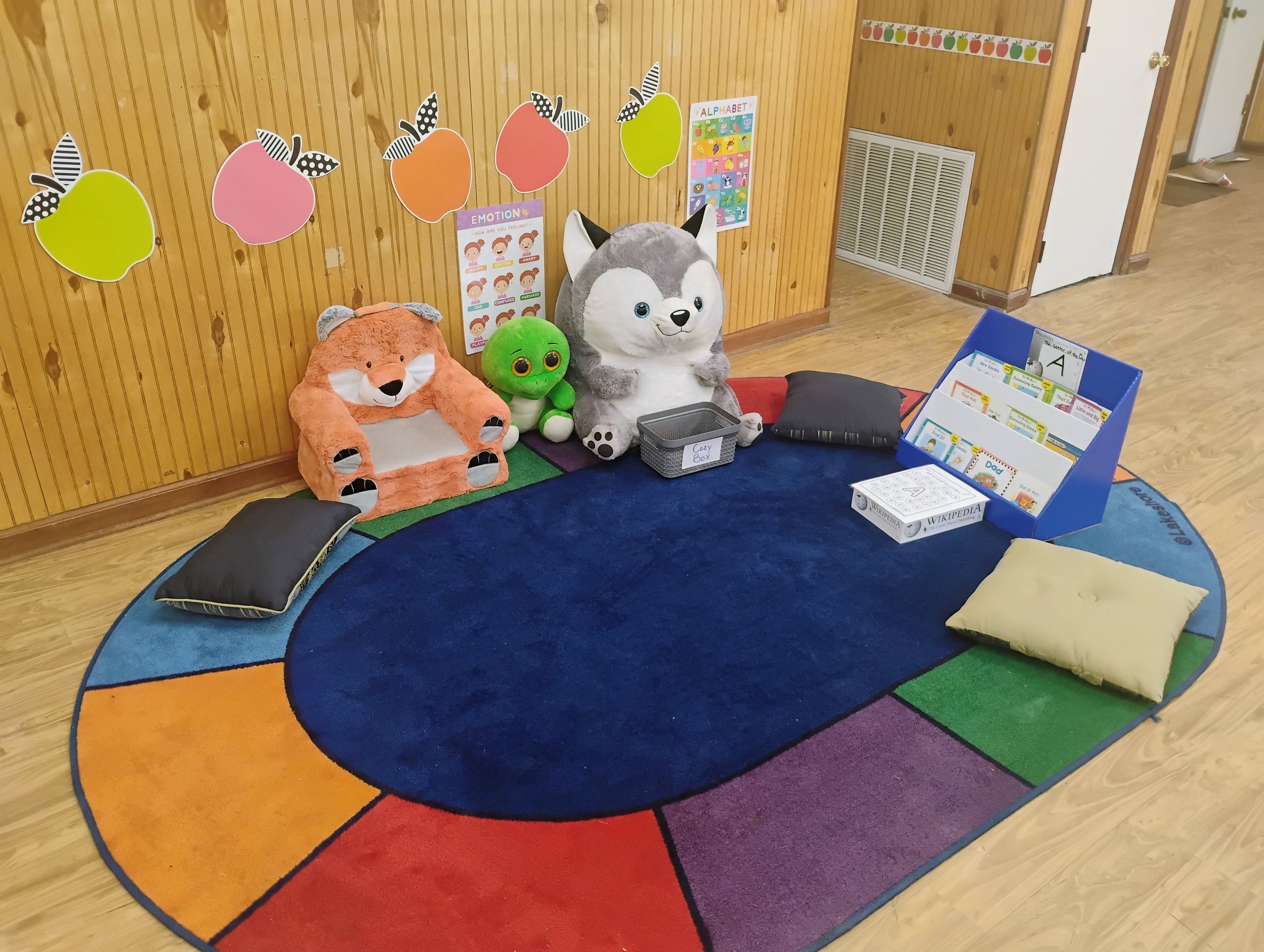 Colorful circular rug with rainbow segments around its edge, surrounded by cushions and stuffed animals including a fox, a green turtle, and a gray and white husky. There's a wooden wall with bird-shaped wall decorations, an educational poster about 