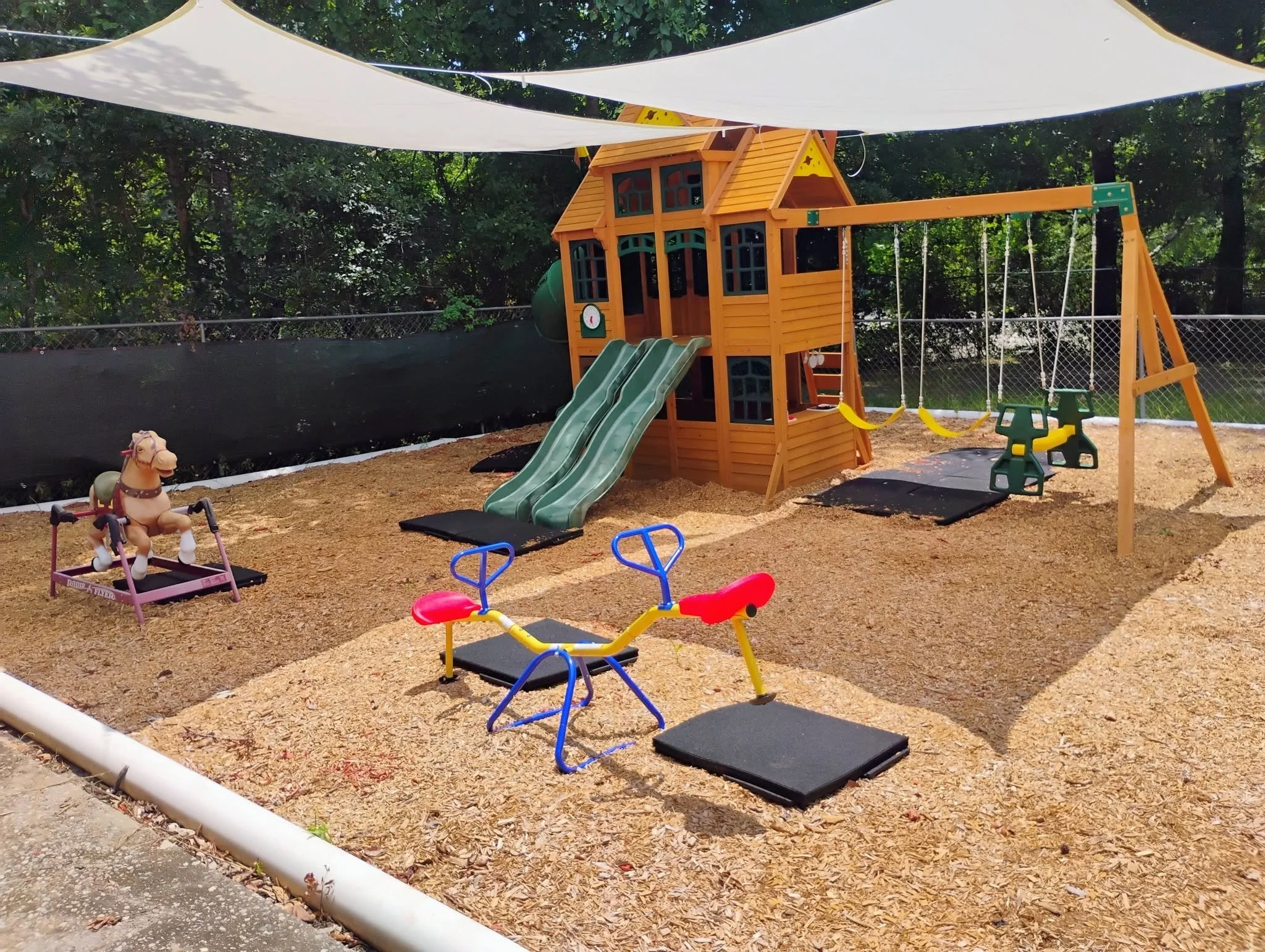 Colorful outdoor playground with slides, swings, and riding toys, surrounded by a safety fence and shaded by trees.