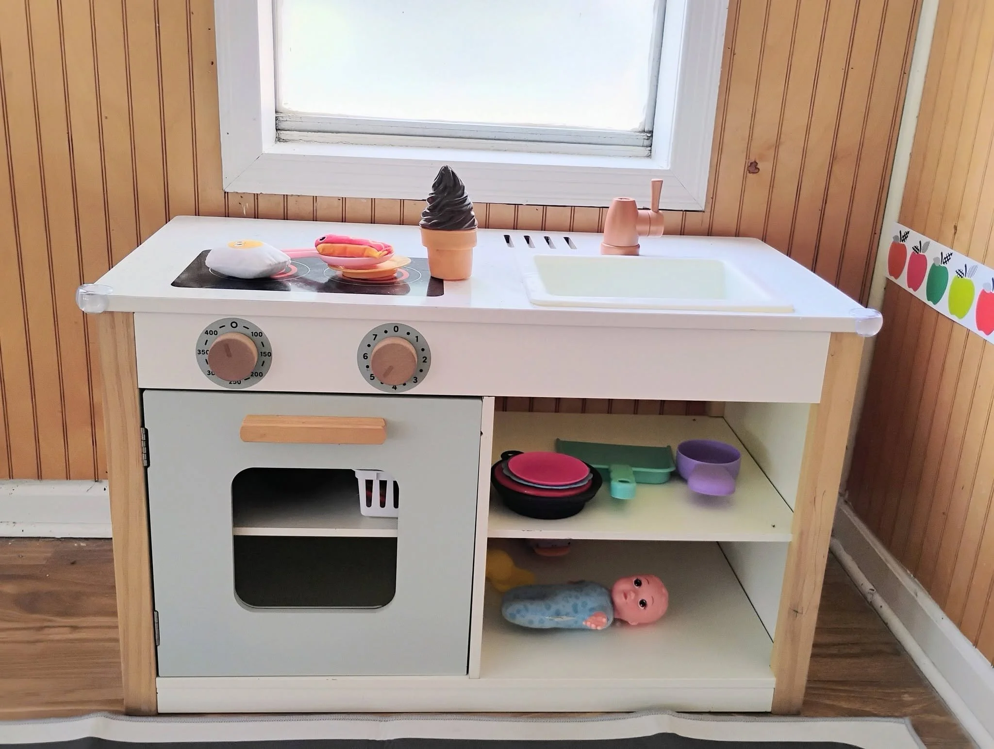 A small white toy kitchen with a stovetop, oven, and sink, decorated with pink, orange, and purple pots and toys, positioned in front of wood-paneled walls and a window.