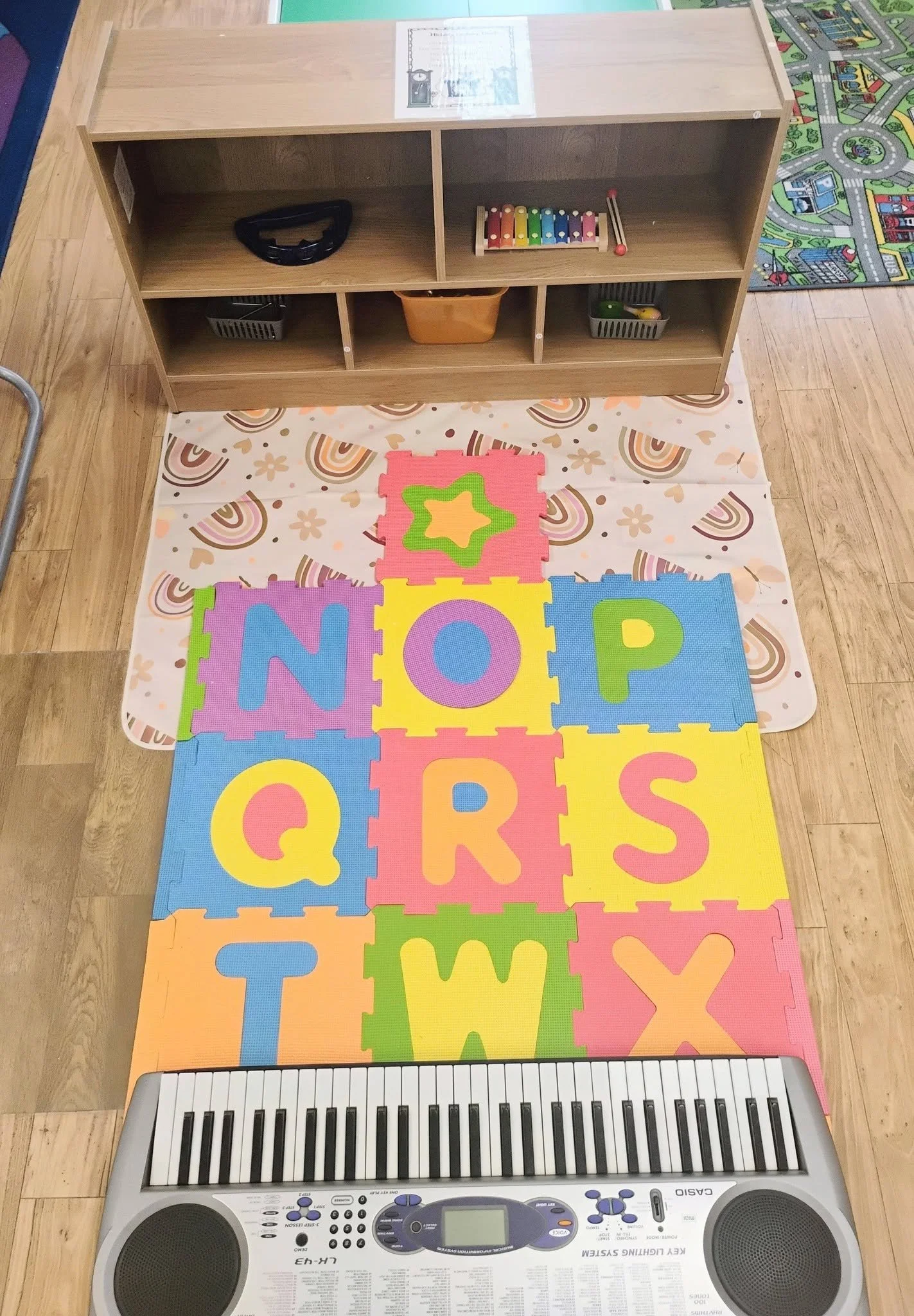 Colorful foam alphabet puzzle mat on a wooden floor, with a keyboard at the bottom and a wooden shelf with children’s toys in the background.