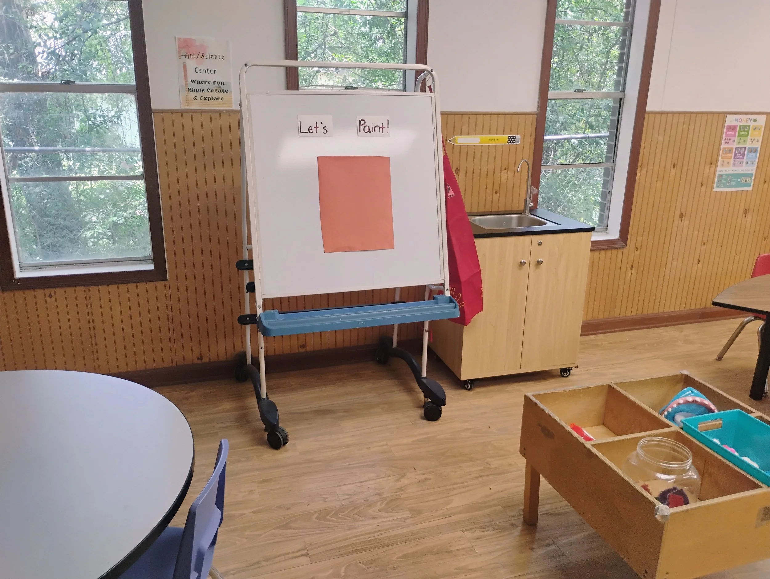 A classroom with large windows overlooking trees. In front, a whiteboard on a stand displays the words 'Let's Paint!' and a piece of orange paper. To the right, there is a small sink, a wooden shelf, and a folder of supplies. Part of a table and chai