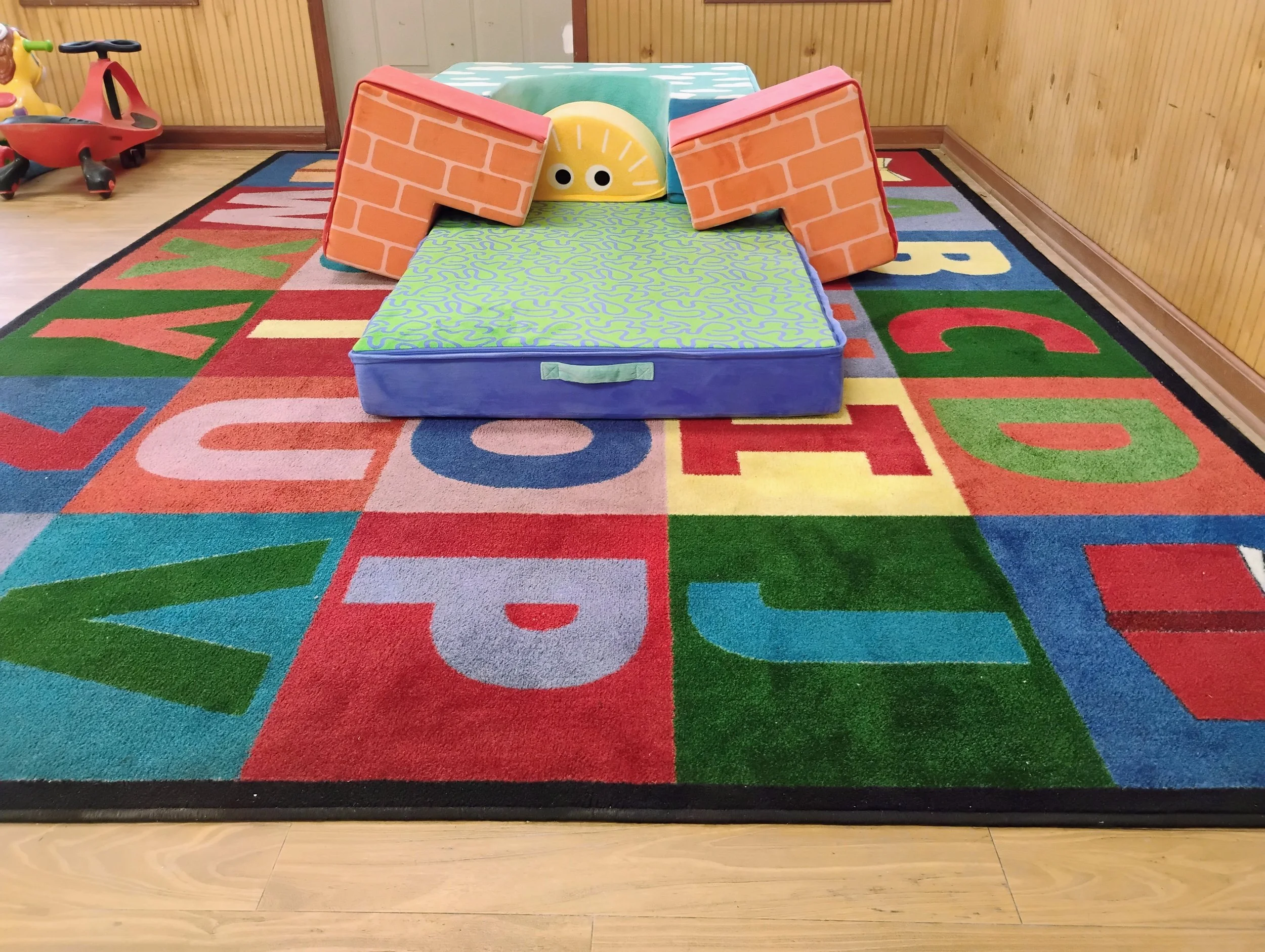 Colorful children's play area with a soft game mat featuring letters, a cardboard tunnel with a face, and a small red ride-on toy in the background.