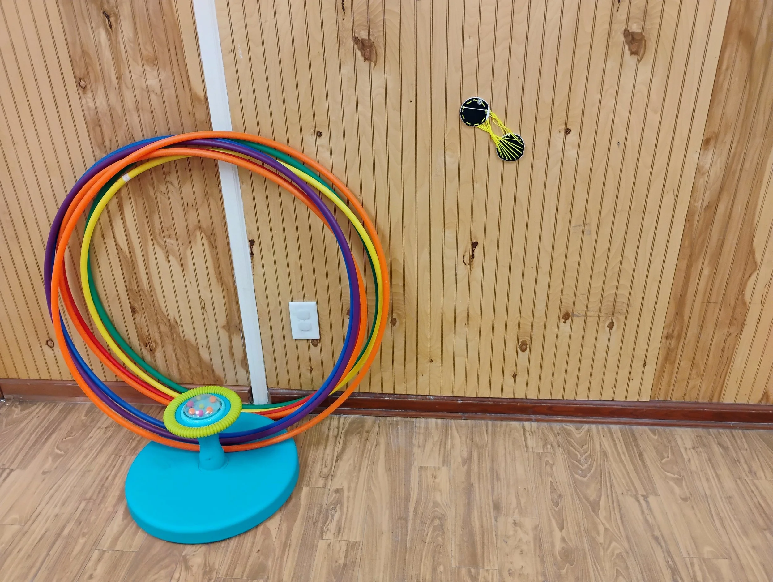Colorful indoor theater or game display with hula hoops arranged on a stand and two beaded ball toys hanging on the wall, set against wooden paneling.