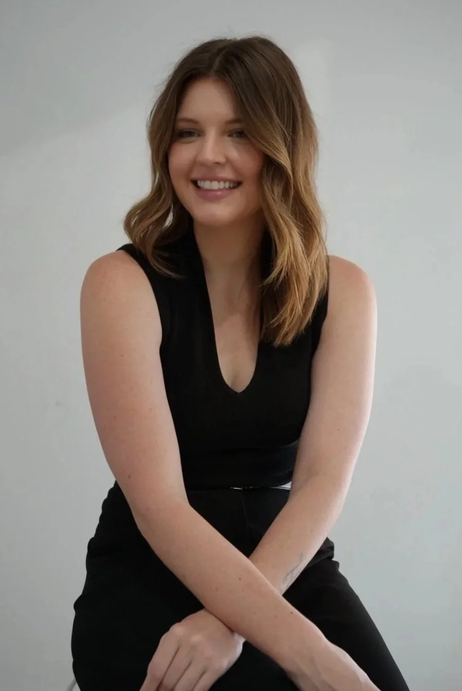 Portrait of a smiling woman with shoulder-length brown hair, wearing a sleeveless black top, sitting against a plain light-colored wall.