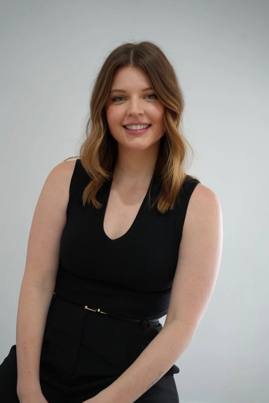 A woman with medium-length brown hair, wearing a sleeveless black top, sitting against a plain white wall, smiling at the camera.