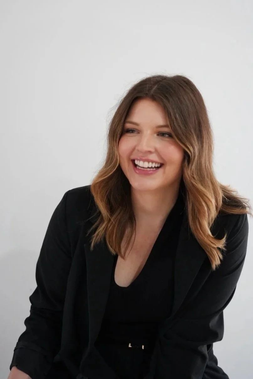 A woman with wavy brown hair smiling and looking to her right, wearing a black blazer and black top, sitting against a plain white background.