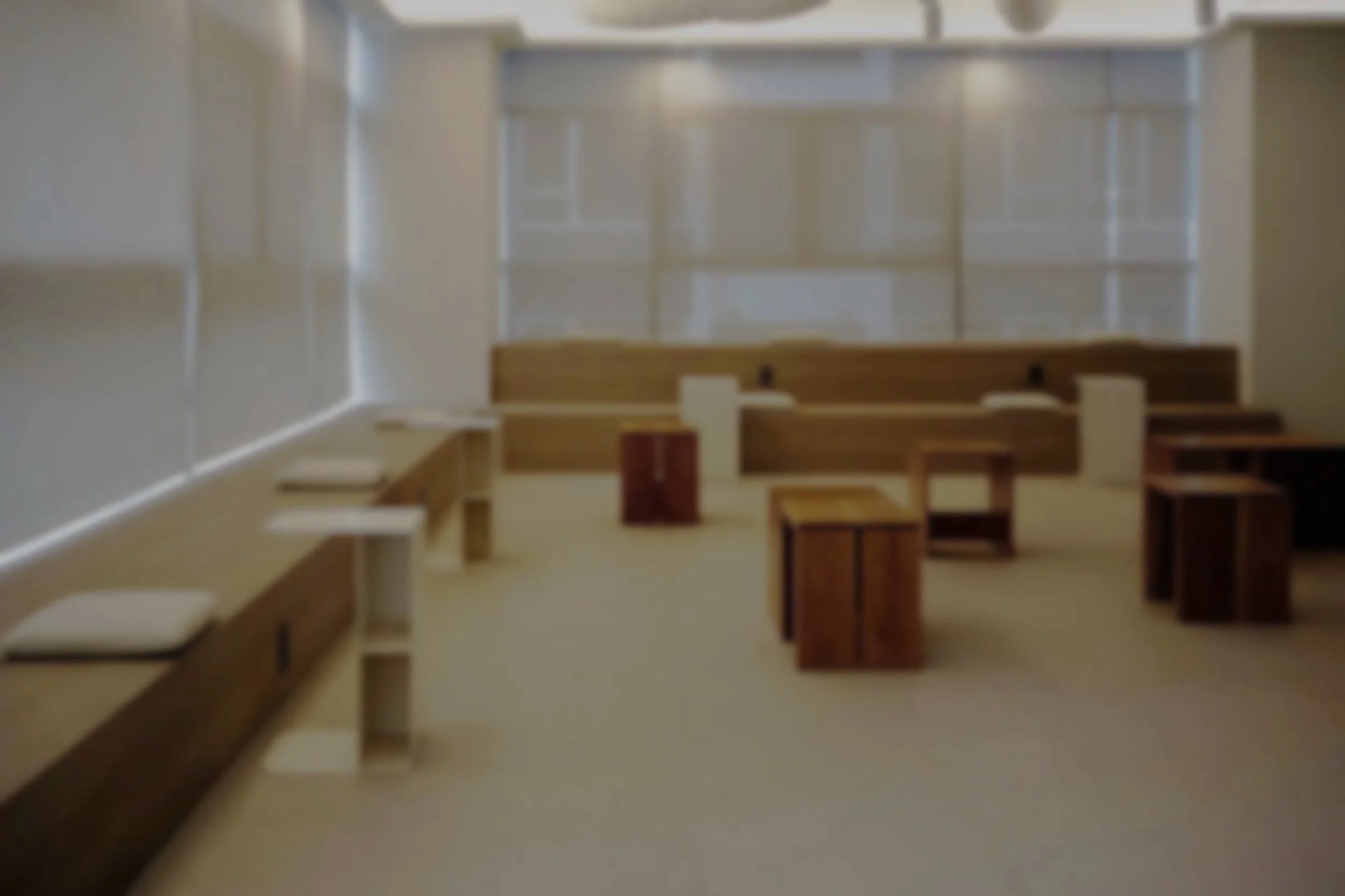 An empty classroom with wooden desks and white walls, with a large window at the back allowing natural light to fill the room.
