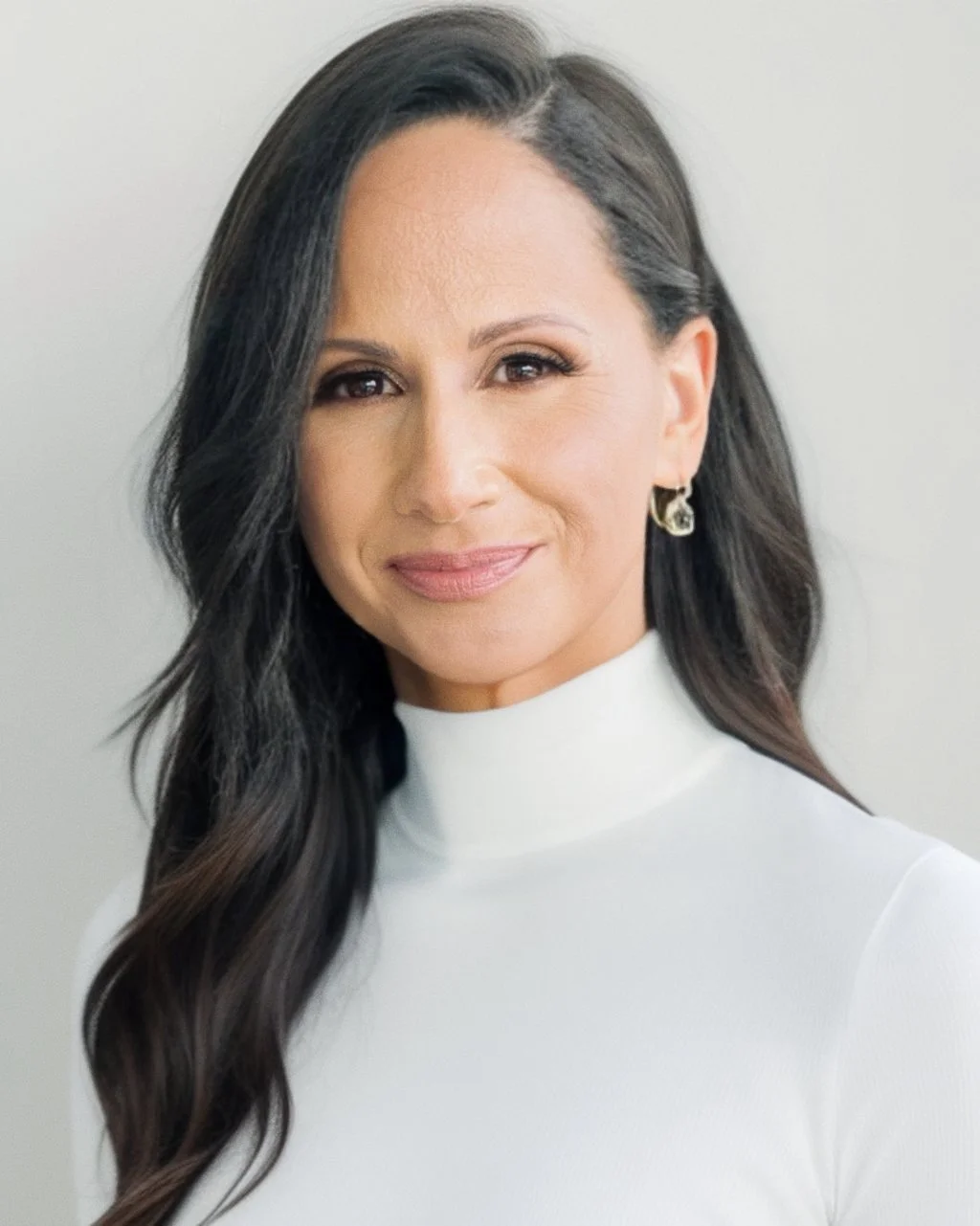 A woman with long, dark hair in loose waves, wearing earrings and a white turtleneck, smiling softly against a plain background.