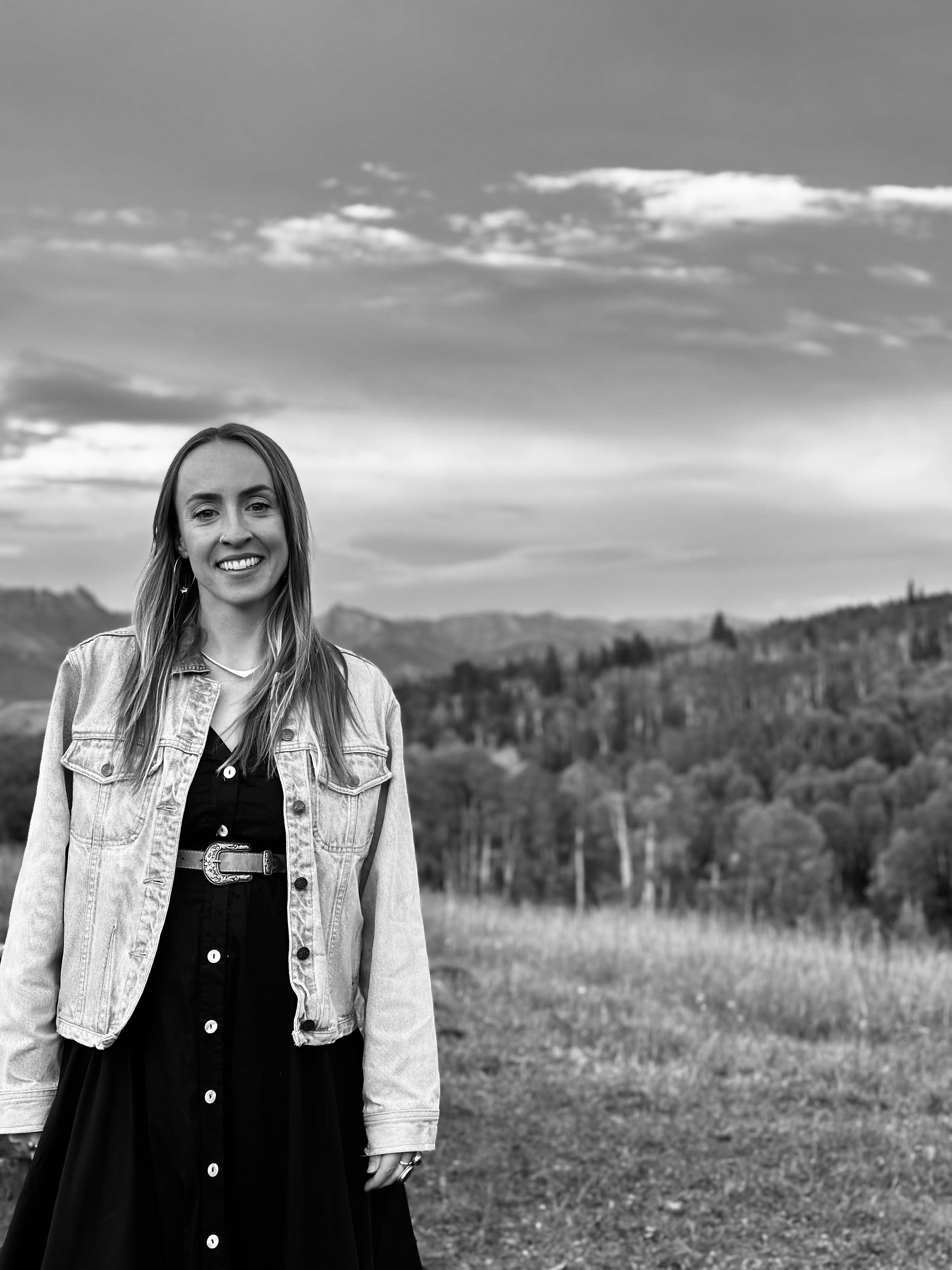 A smiling young woman with long hair, wearing a denim jacket over a black dress with white buttons and a belt, standing outdoors with a scenic landscape of rolling hills and trees in the background under a cloudy sky.