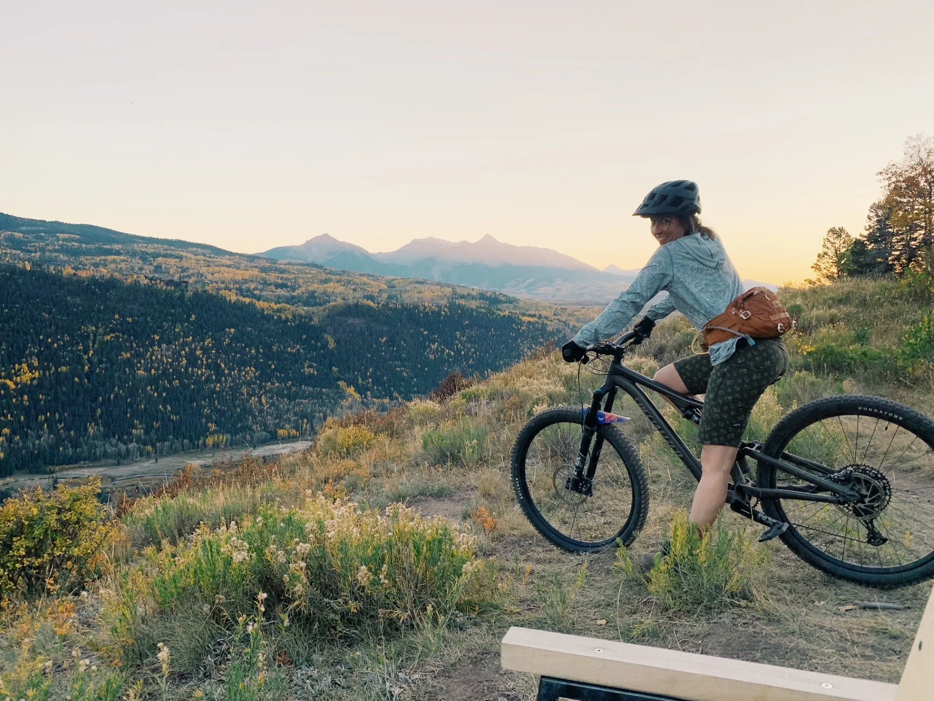 A woman wearing a helmet and athletic clothing riding a mountain bike on a trail with mountains in the background at sunset.