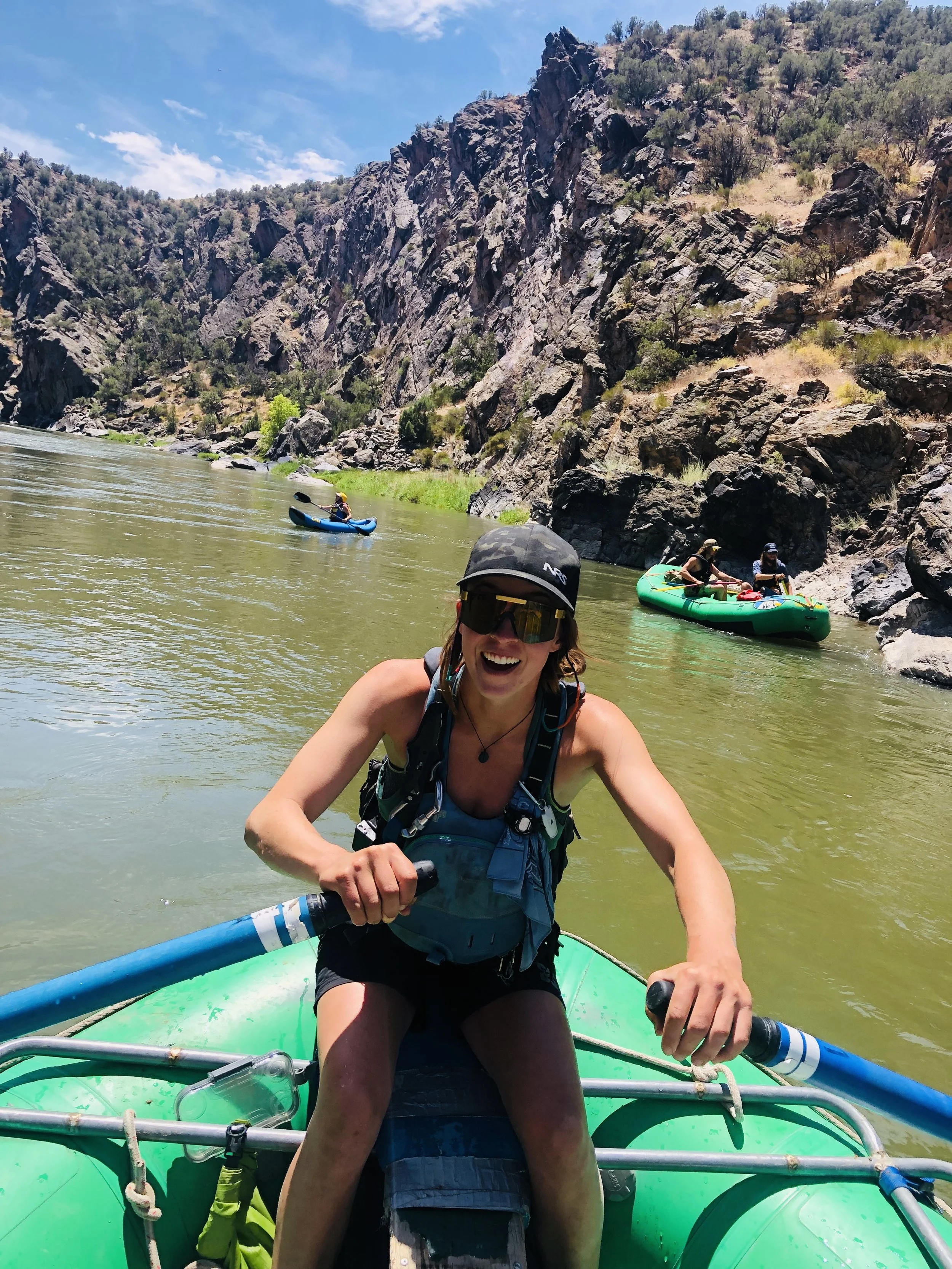 People kayaking on a river with rocky cliffs and lush greenery in the background, smiling and enjoying outdoor water activities.