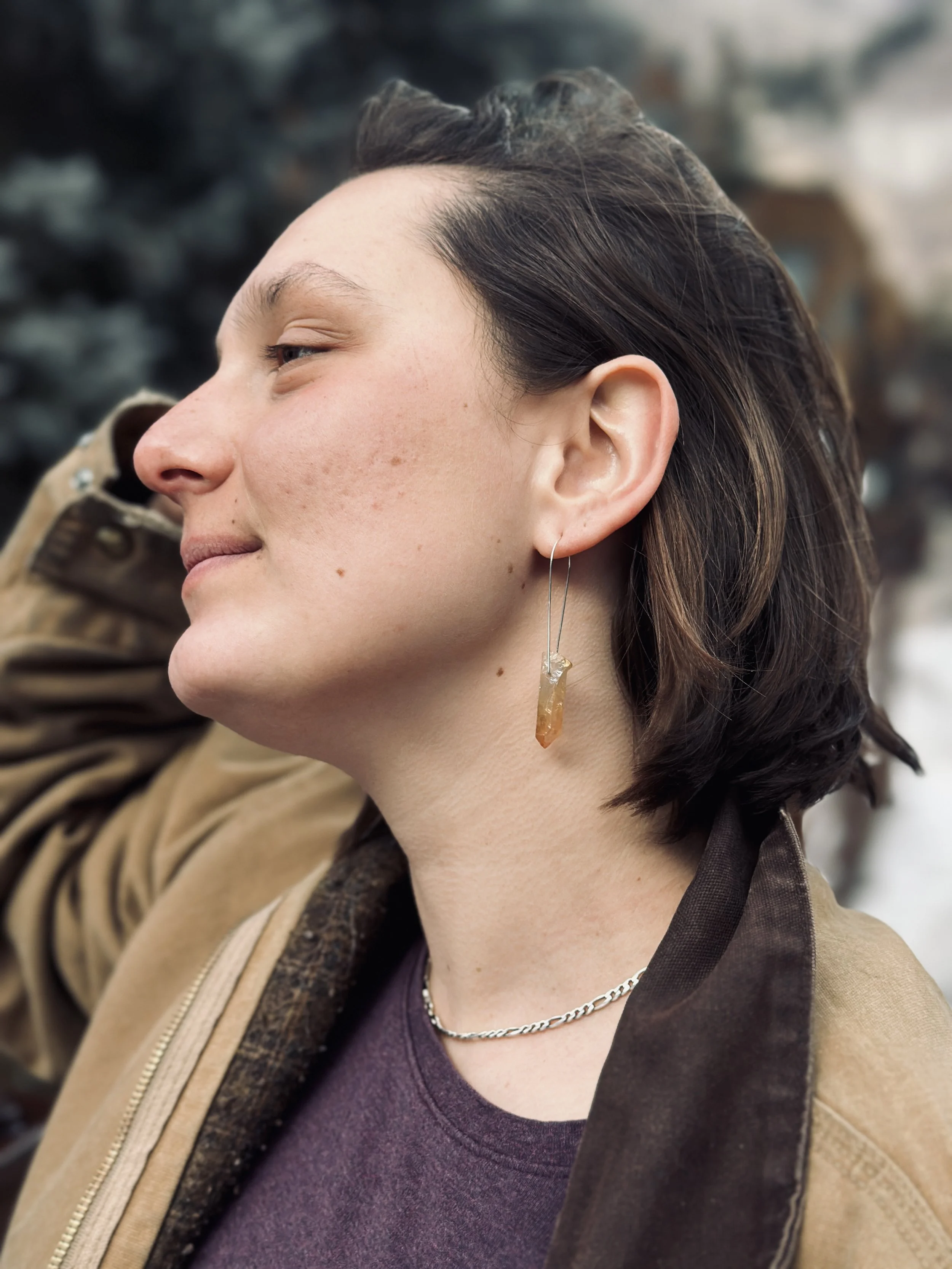 Close-up of a woman from the side, smiling, with earrings, necklace, and wearing a jacket.