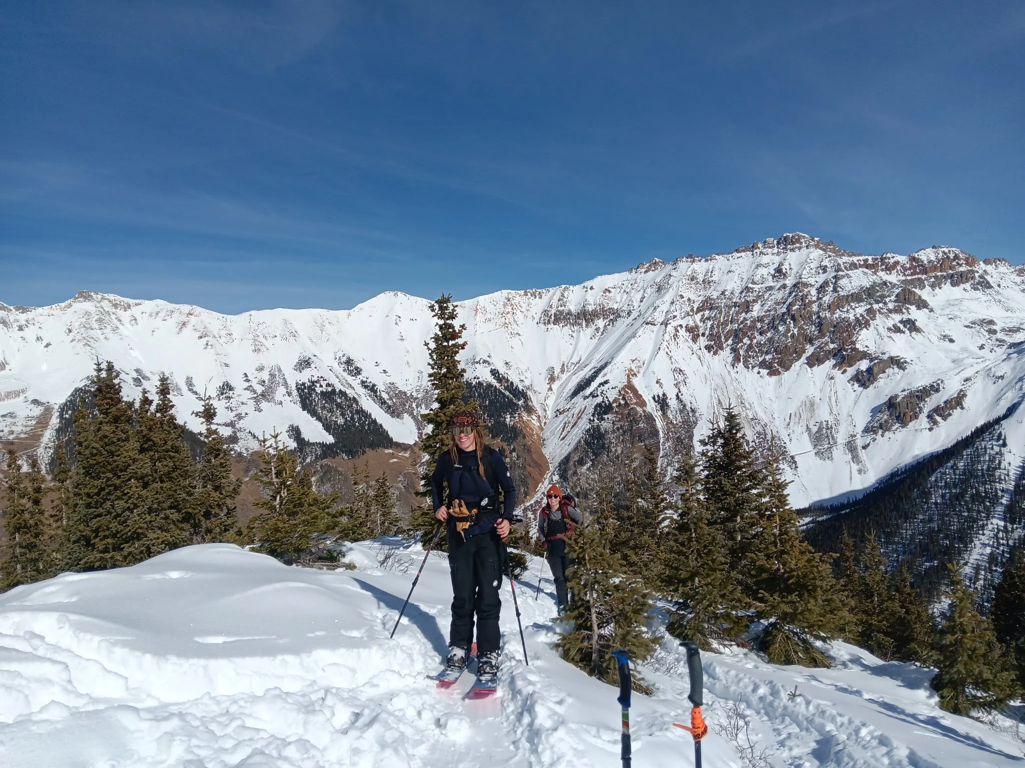 Two women dressed in winter gear, standing in snow with ski poles, surrounded by pine trees and a snow-covered mountain range under a clear blue sky.